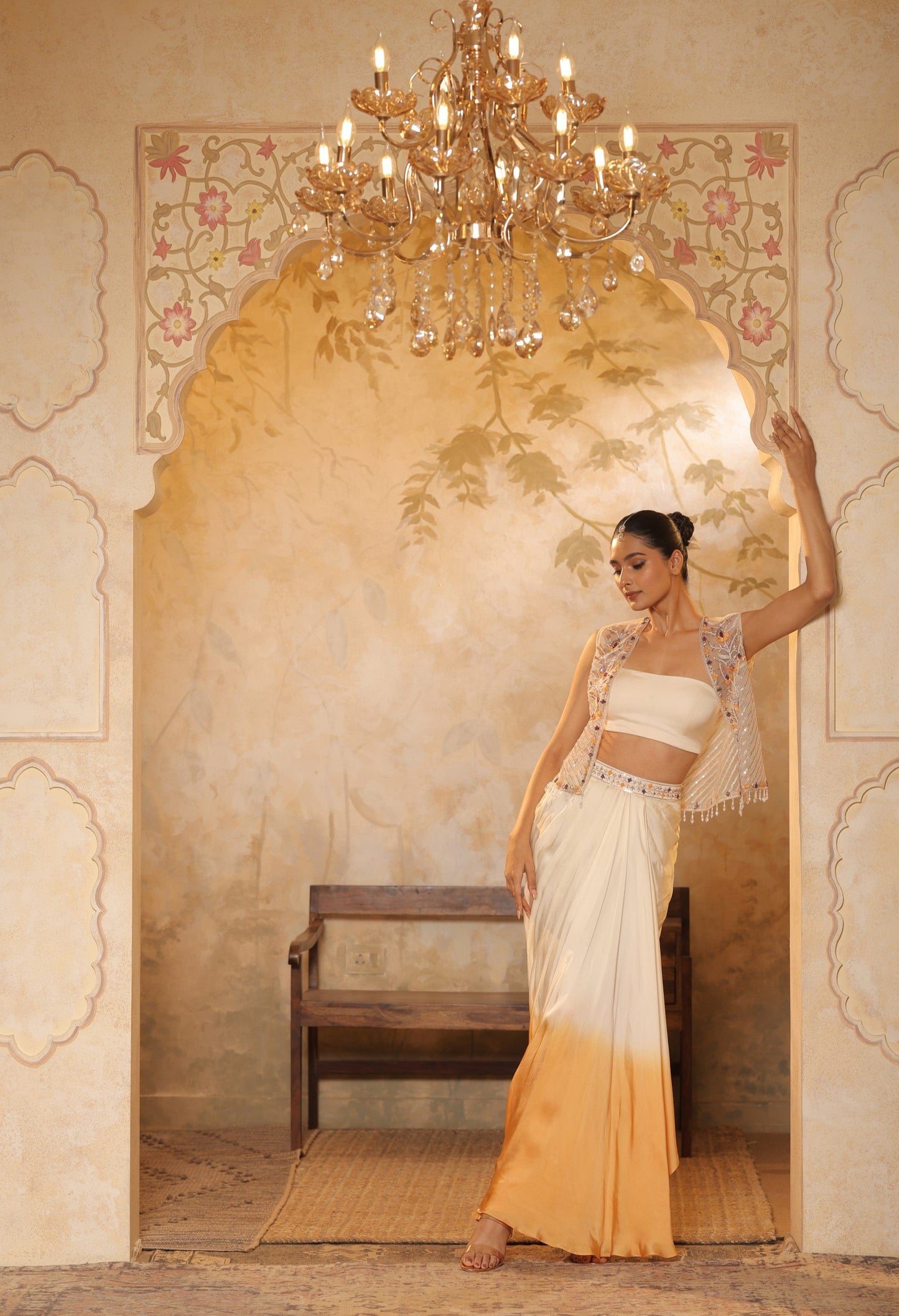 Woman in a white and yellow saree standing in a decorated room with a chandelier. Ivory orange skirt dress, hand embroidered crepe outfit, elegant festive wear, designer ethnic attire, premium traditional ensemble, Sunanta Madaan, near me, Mumbai, Delhi, Hyderabad, Bangalore, Pune