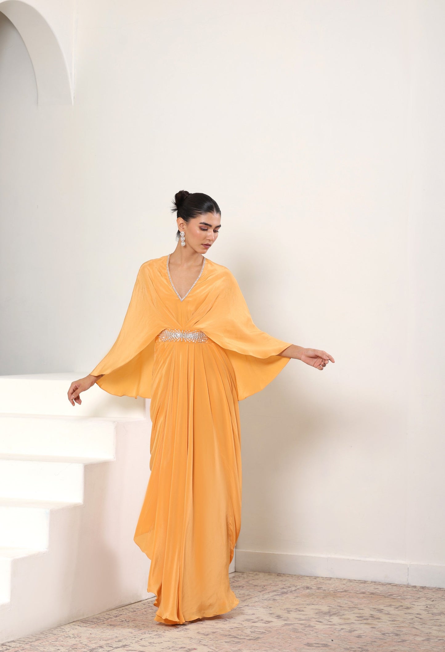 Woman in a yellow evening gown standing on a staircase with a white wall background; Orange crepe kaftan, embroidered designer kaftan, luxury evening wear, elegant festive outfit, premium ethnic kaftan, Sunanta Madaan