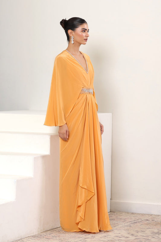 Woman in a long orange dress standing on a staircase with a white wall background; Orange crepe kaftan, embroidered designer kaftan, luxury evening wear, elegant festive outfit, premium ethnic kaftan, Sunanta Madaan