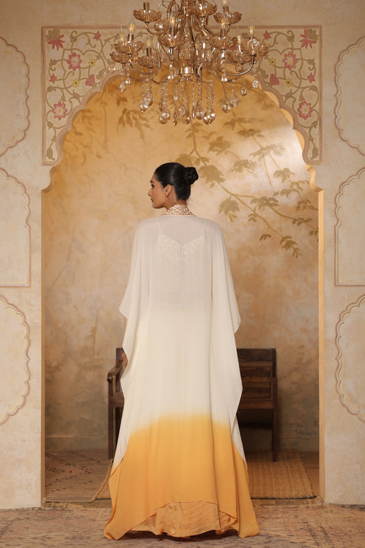 Woman in a white and yellow dress standing in a decorated room with a chandelier. Ivory orange organza lehenga, satin embroidered lehenga set, festive lehenga with cape, elegant designer outfit, ethnic party wear, Sunanta Madaan