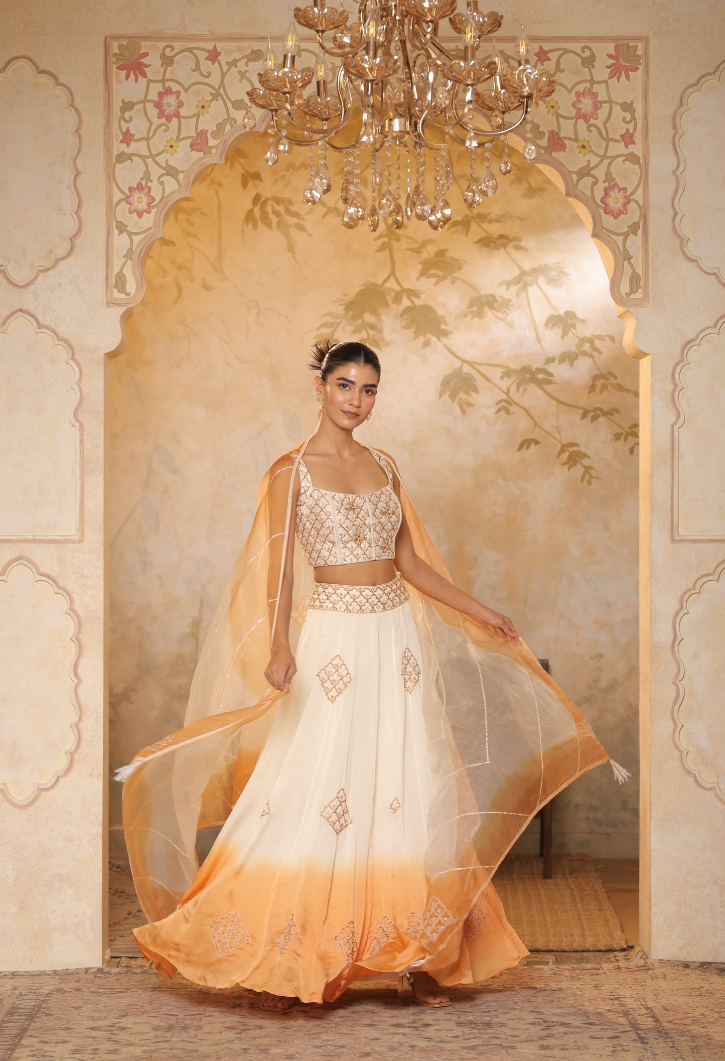 Woman in a white and orange traditional outfit standing in a decorated room with a chandelier. Sunanta Madaan – Ivory-Orange Satin Organza Lehenga, Embroidered Designer Outfit, Elegant Festive Attire, Premium Party & Reception Wear, Boho Indo-Western Dress, Indo-Western Fusion Wear, Indo-Western Attire for Engagement, Indo-Western Outfit for Sangeet, Indo-Western Draped & Embroidered Skirt, Mumbai, Delhi, Hyderabad, Bangalore, Pune, near me