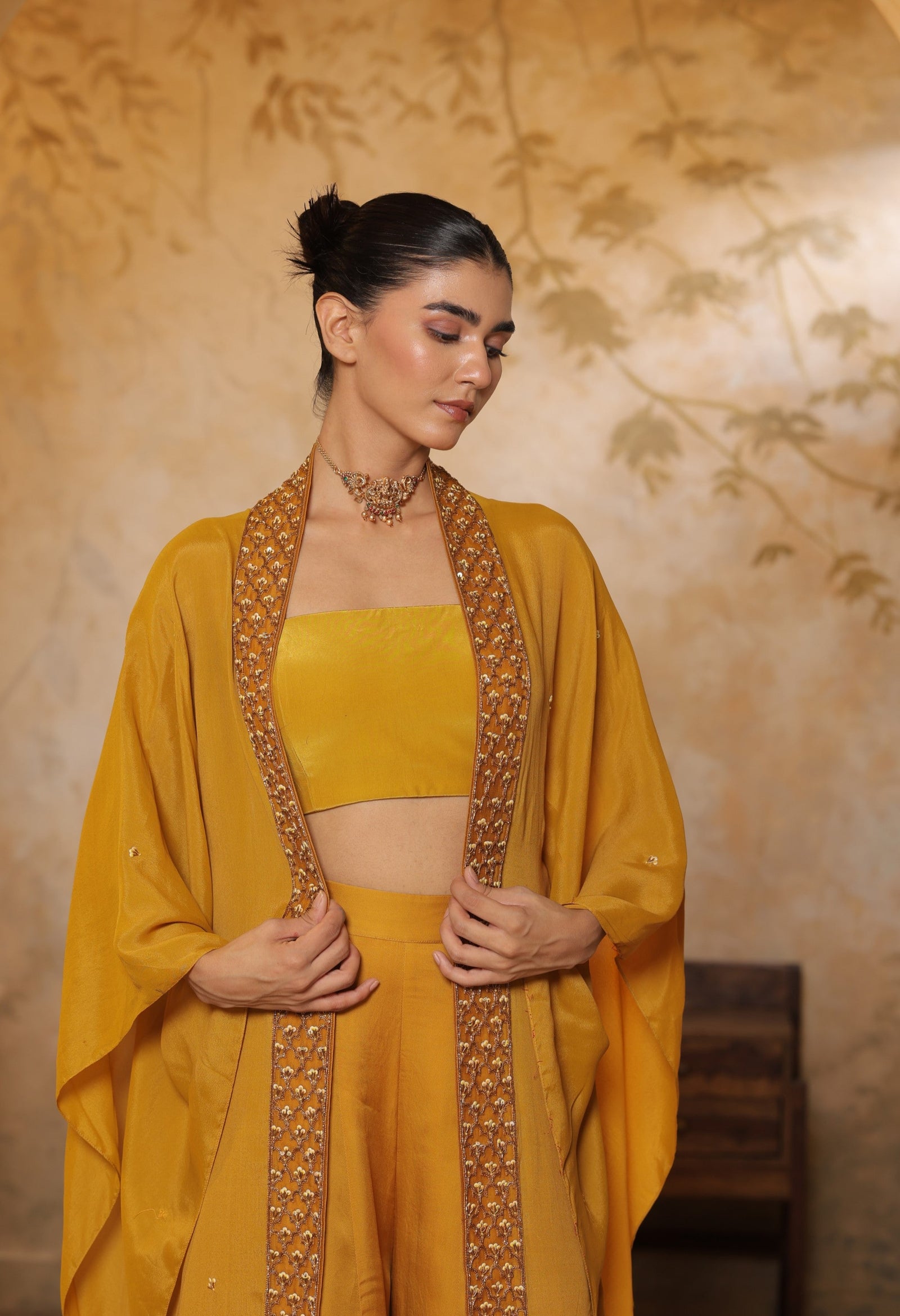 Woman wearing a mustard yellow traditional outfit with intricate designs against a floral-patterned wall.