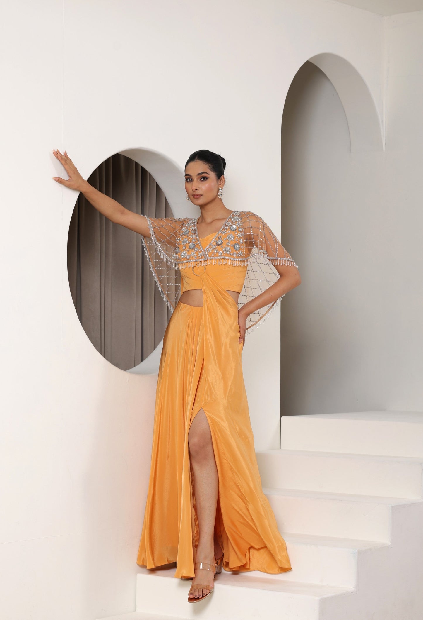 Woman in a bright orange saree with sheer overlay standing on a staircase. premium evening wear, Orange crepe saree, embroidered cape saree, designer reception saree, elegant festive outfit, Sunanta Madaan