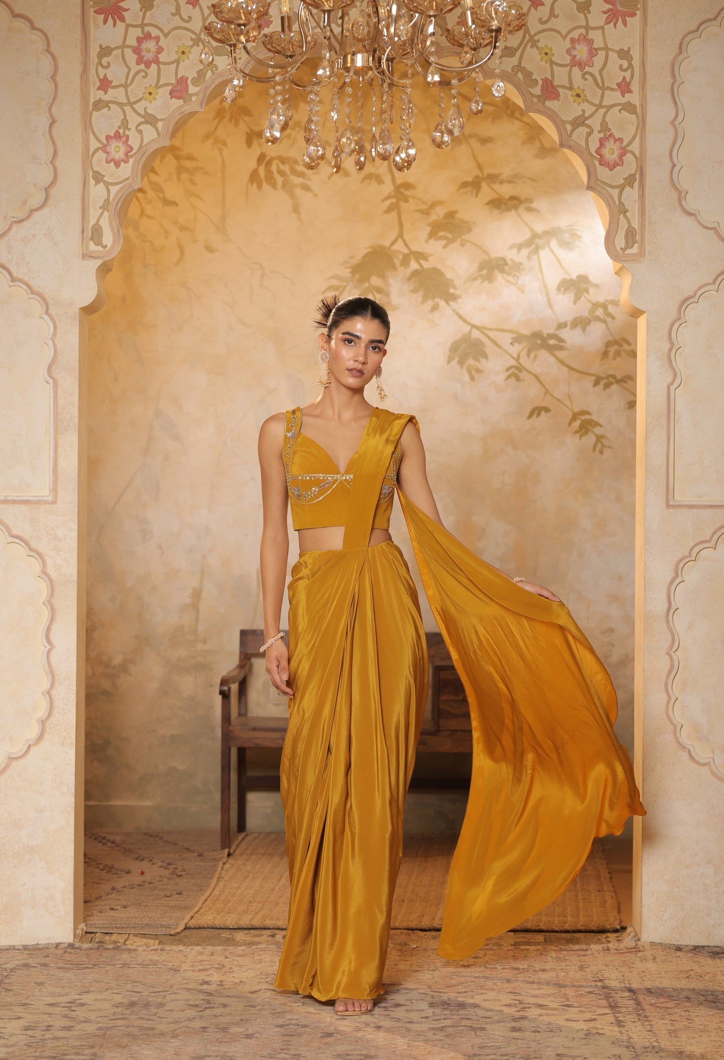 Woman in a yellow saree standing in an ornate room with a chandelier. Golden yellow crepe saree, haldi ceremony saree, embroidered festive saree, designer ethnic wear, elegant haldi outfit, Sunanta Madaan