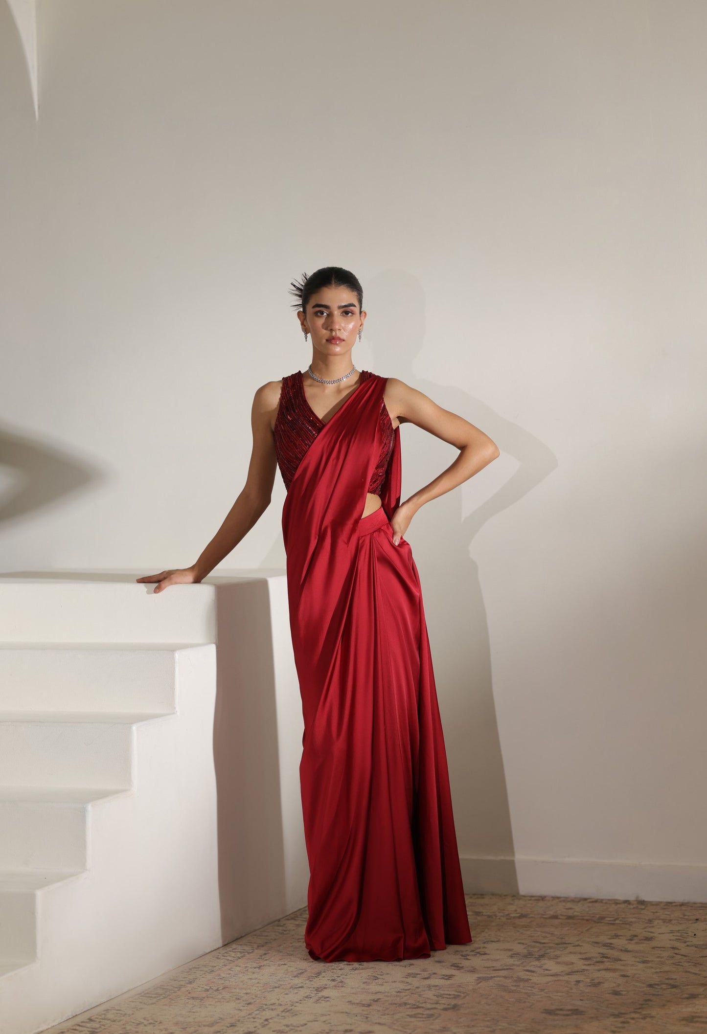 Woman in a red evening gown standing on a staircase with a plain wall background; Sunanta Madaan – Maroon Satin Embroidered Draped Saree, Elegant Wedding Wear, Premium Festive Outfit, Boho Indo-Western Dress, Indo-Western Fusion Wear, Indo-Western Attire for Engagement, Indo-Western Outfit for Sangeet, Indo-Western Draped & Embroidered Saree, Women’s Designer Bridal Ensemble, Delhi, Jaipur, Hyderabad, Ahmedabad, Bangalore, near me