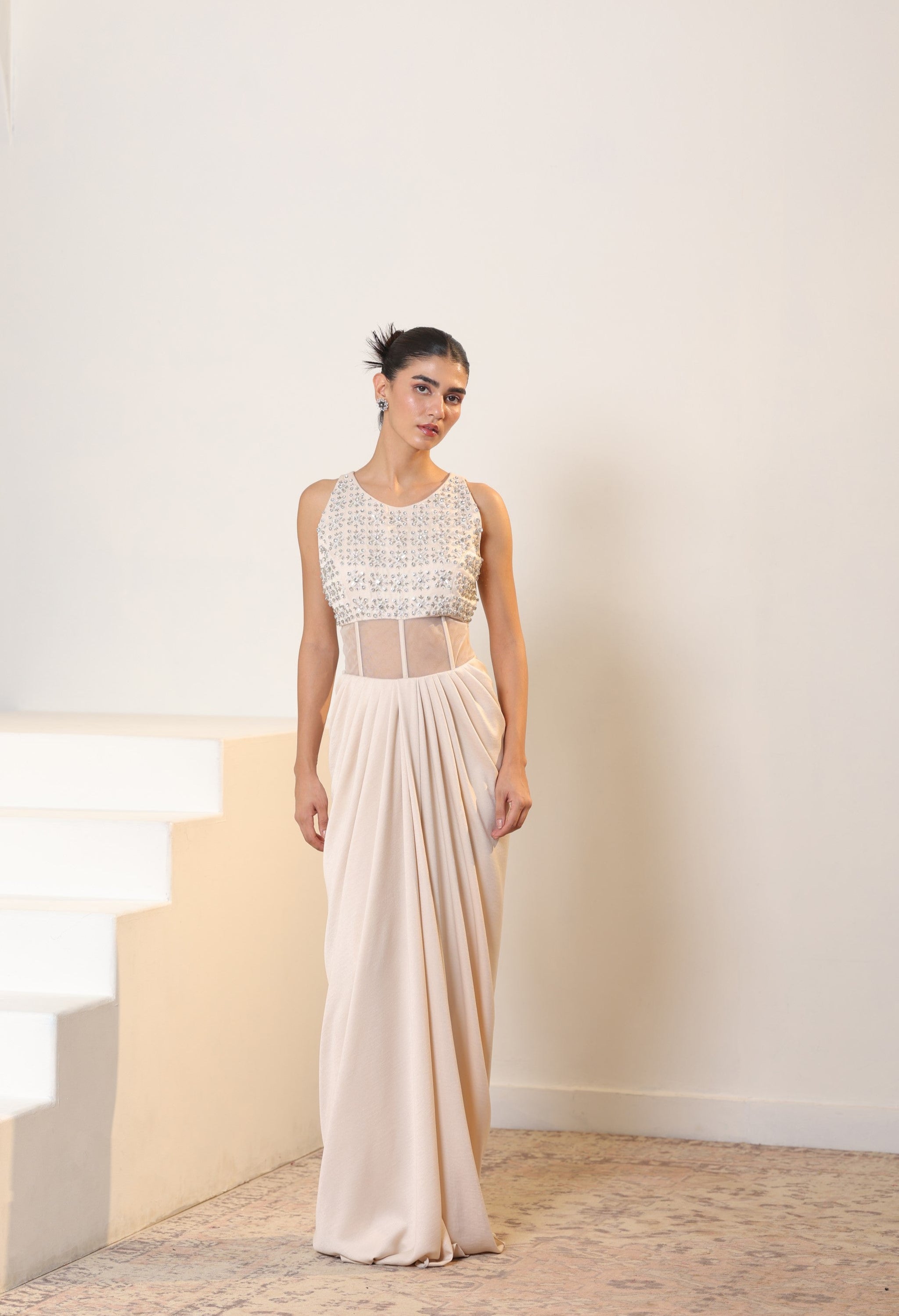 Woman wearing a white lace top and beige skirt standing on a staircase. Sunanta Madaan – Ivory Crepe Satin Western Embroidered Gown, Premium Cocktail Wear, Elegant Party Outfit, Boho Indo-Western Dress, Indo-Western Fusion Wear, Women’s Formal Evening Dress, Indo-Western Outfit for Reception, Party Outfit, Indo-Western Draped Dress, Jaipur, Delhi, Hyderabad, Ahmedabad, Bangalore, near me
