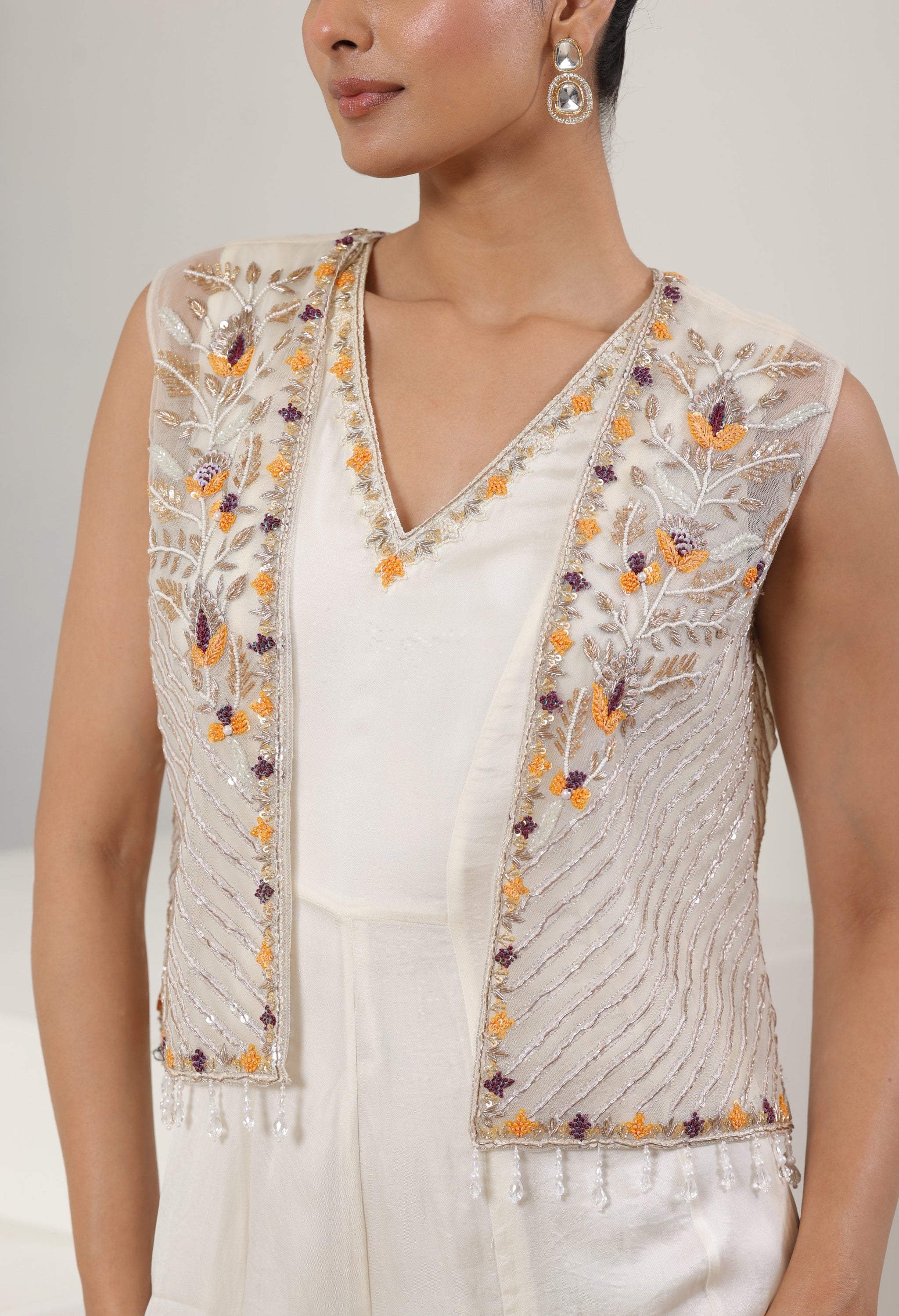 Woman wearing a white embroidered vest with orange and purple details on a plain background; Ivory orange jumpsuit, satin organza outfit, designer cape jumpsuit, elegant occasional wear, festive ethnic attire, Sunanta Madaan, near me, Mumbai, Delhi, Hyderabad, Bangalore, Pune