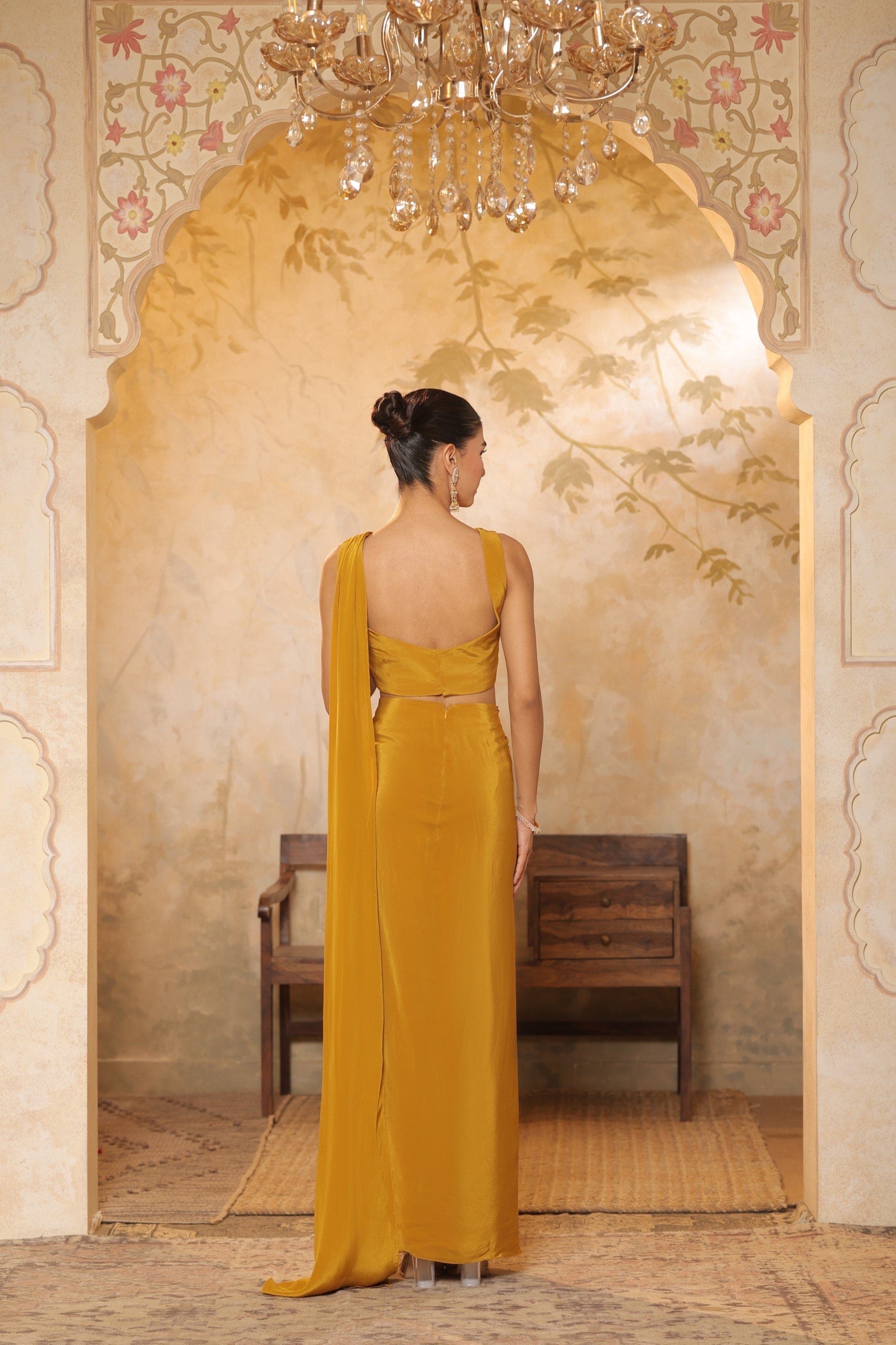 Woman in a yellow saree standing in an ornate room with a chandelier. Golden yellow crepe saree, haldi ceremony saree, embroidered festive saree, designer ethnic wear, elegant haldi outfit, Sunanta Madaan