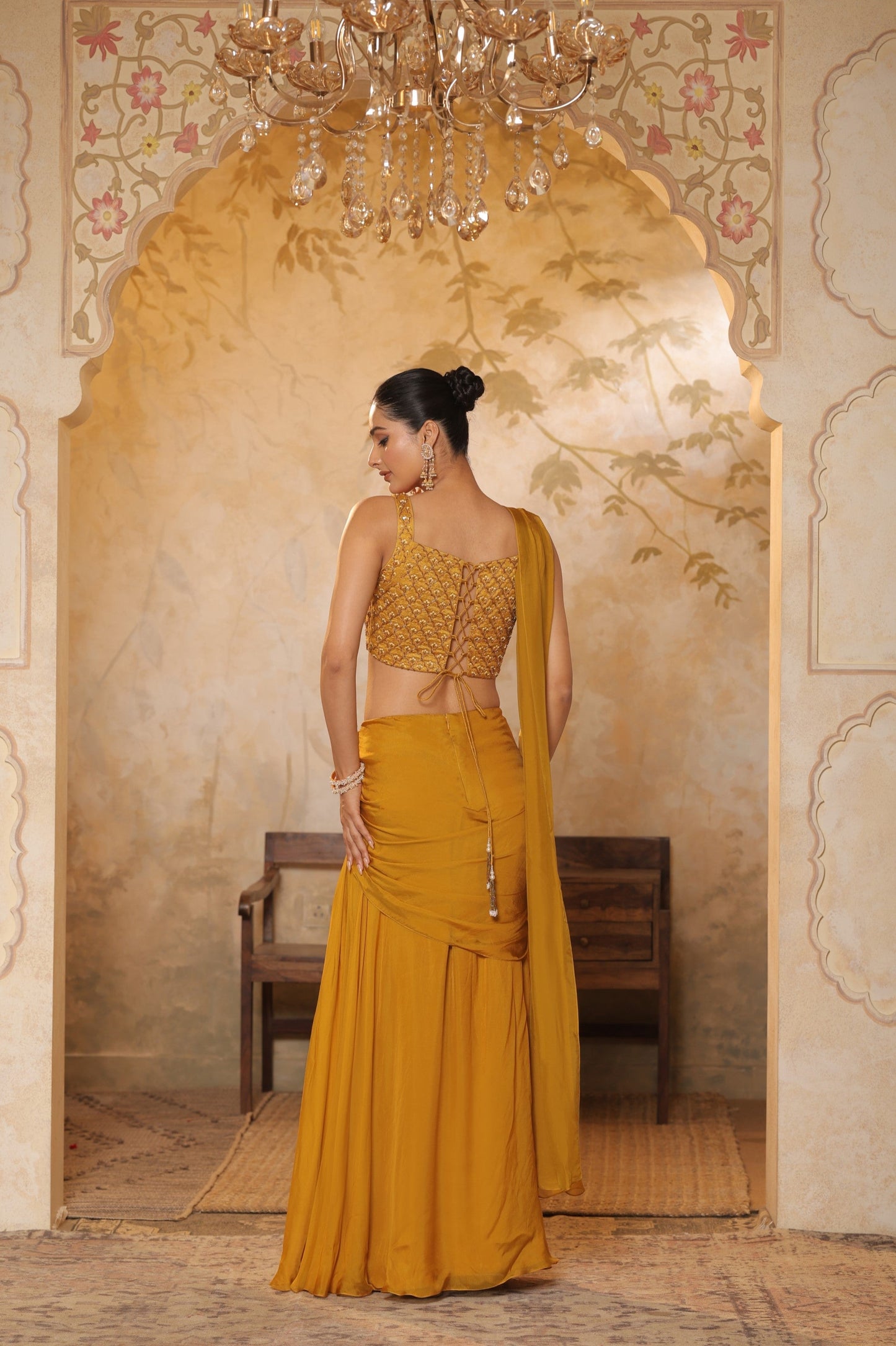Woman in a mustard yellow traditional outfit standing in an ornate room with a chandelier. Golden yellow saree, chiffon embroidered saree, elegant traditional saree, festive ethnic wear, designer saree online, Sunanta Madaan