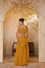 Woman in a mustard yellow traditional outfit standing in an ornate room with a chandelier. Golden yellow saree, chiffon embroidered saree, elegant traditional saree, festive ethnic wear, designer saree online, Sunanta Madaan