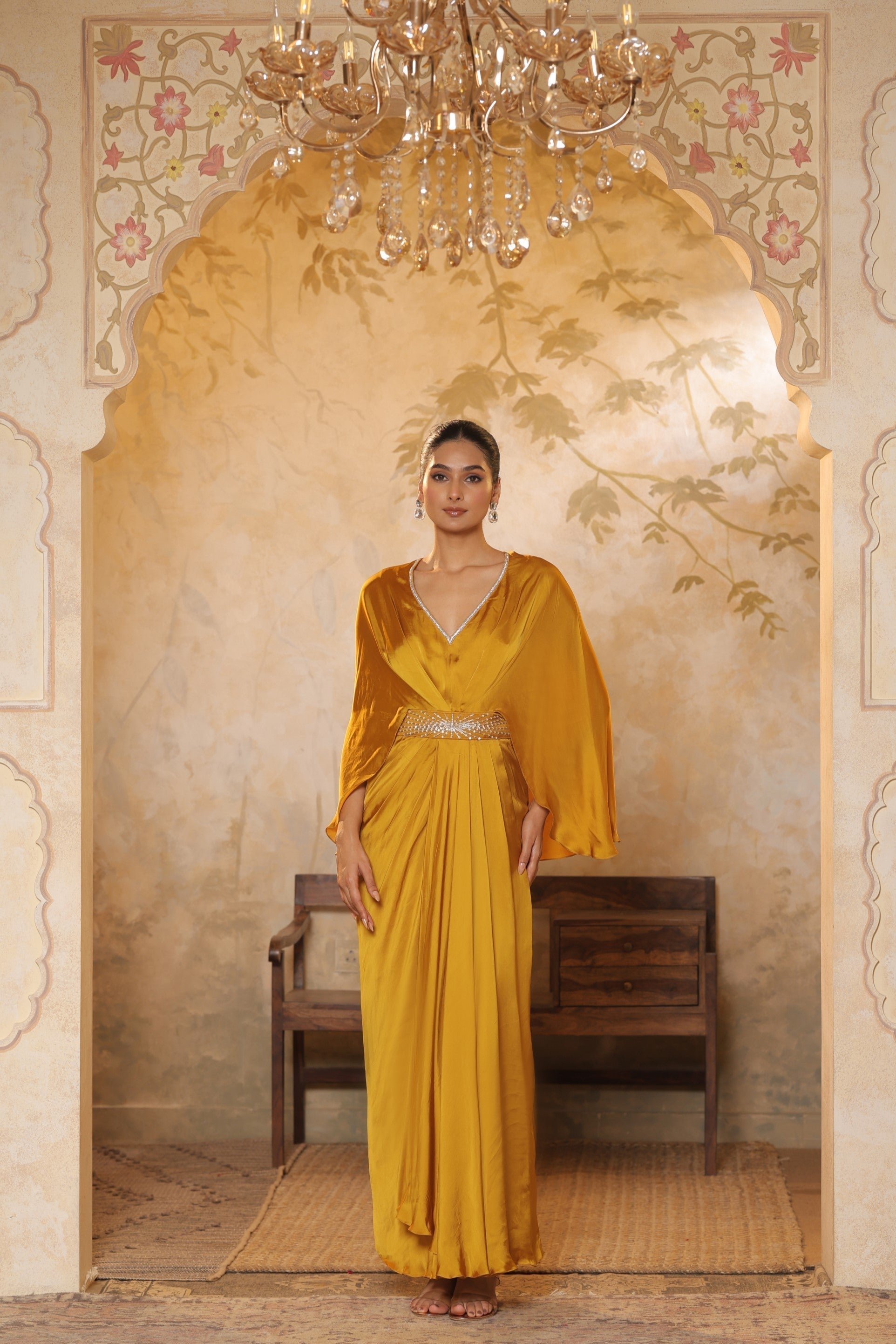 Woman in a yellow dress standing in an ornate room with a chandelier. Yellow crepe kaftan, V-neck embroidered kaftan, handcrafted festive outfit, elegant ethnic wear, designer party kaftan, Sunanta Madaan