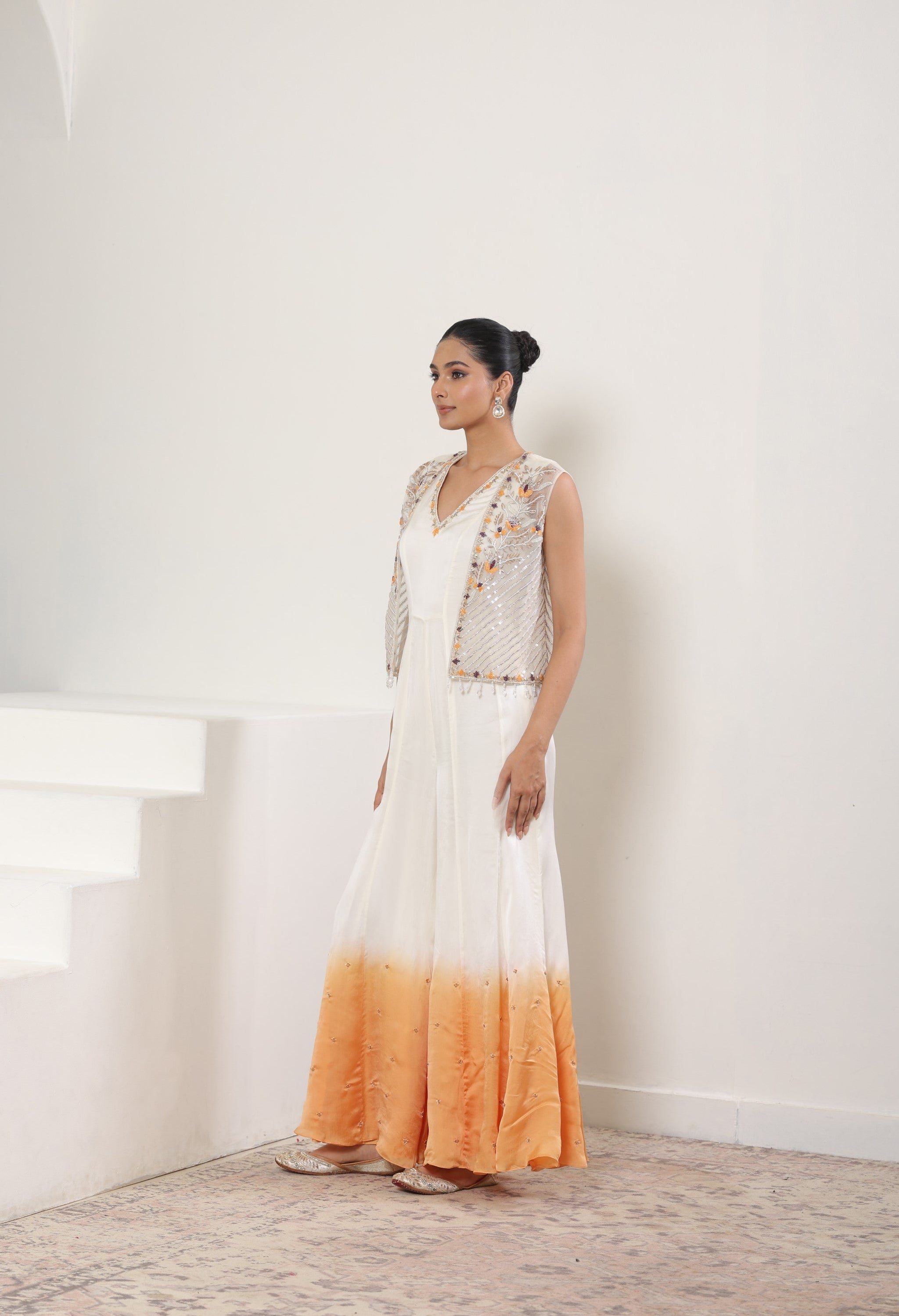 Woman wearing a long, gradient dress with a white top and orange bottom in a minimalistic setting. Ivory orange jumpsuit, satin organza outfit, designer cape jumpsuit, elegant occasional wear, festive ethnic attire, Sunanta Madaan, near me, Mumbai, Delhi, Hyderabad, Bangalore, Pune