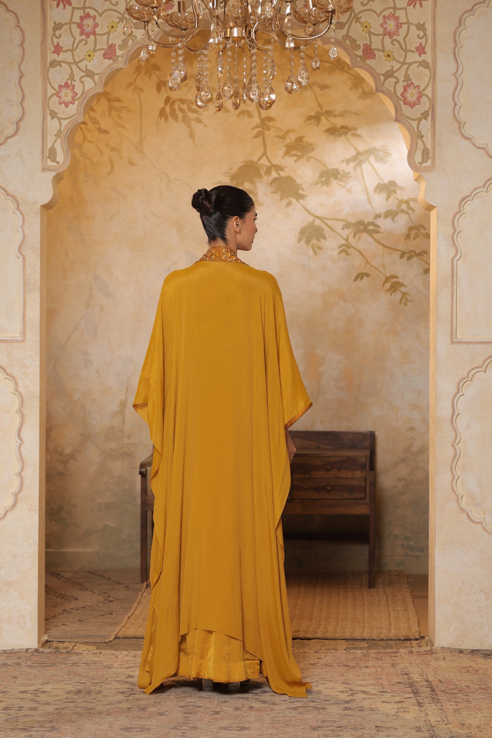 Woman in a yellow dress standing in an ornate room with a chandelier. Golden yellow sharara set, embroidered wedding outfit, designer festive wear, elegant ethnic ensemble, premium traditional attire, Sunanta Madaan