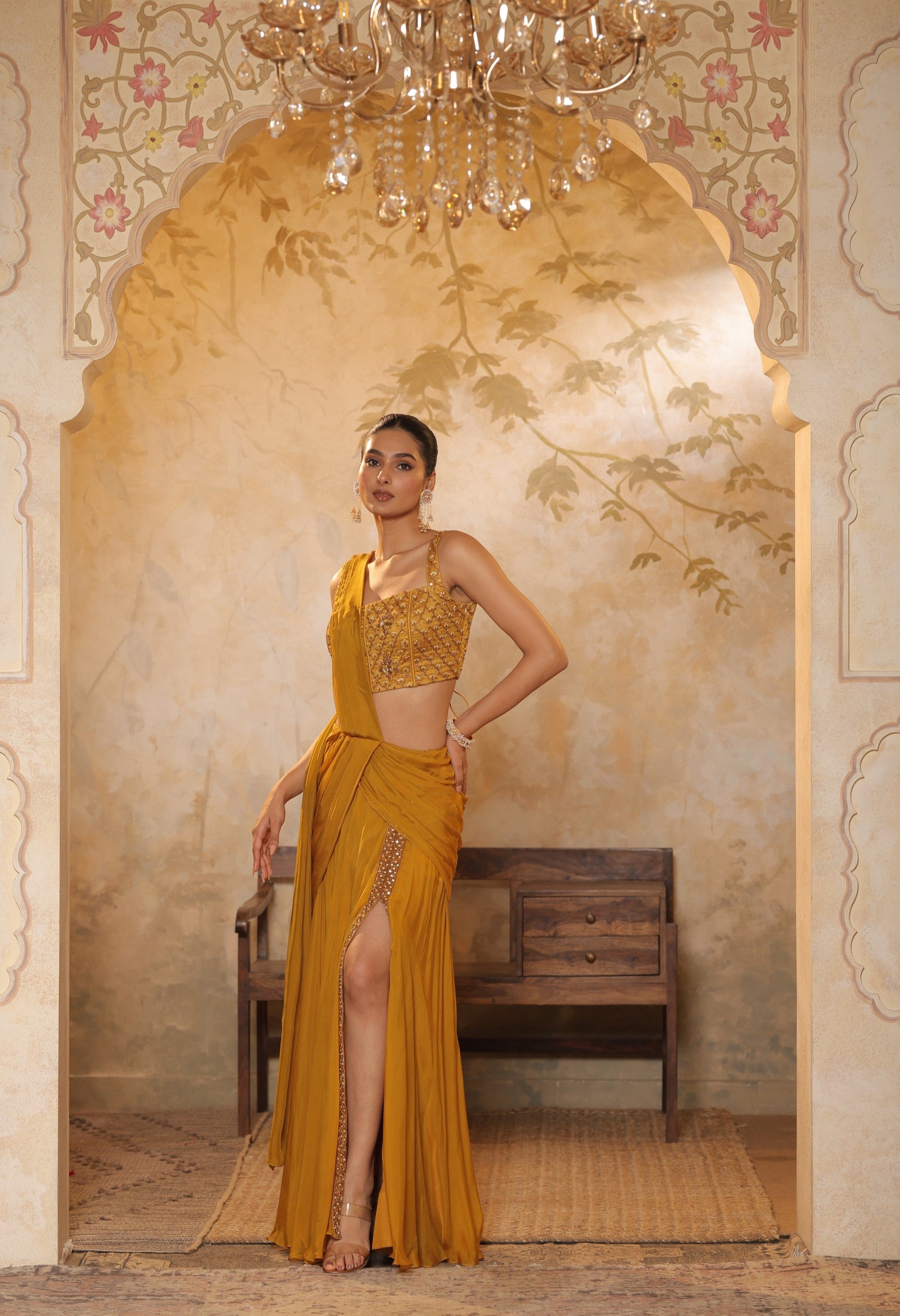Woman in a mustard yellow traditional outfit standing in an ornate doorway. Golden yellow saree, chiffon embroidered saree, elegant traditional saree, festive ethnic wear, designer saree online, Sunanta Madaan