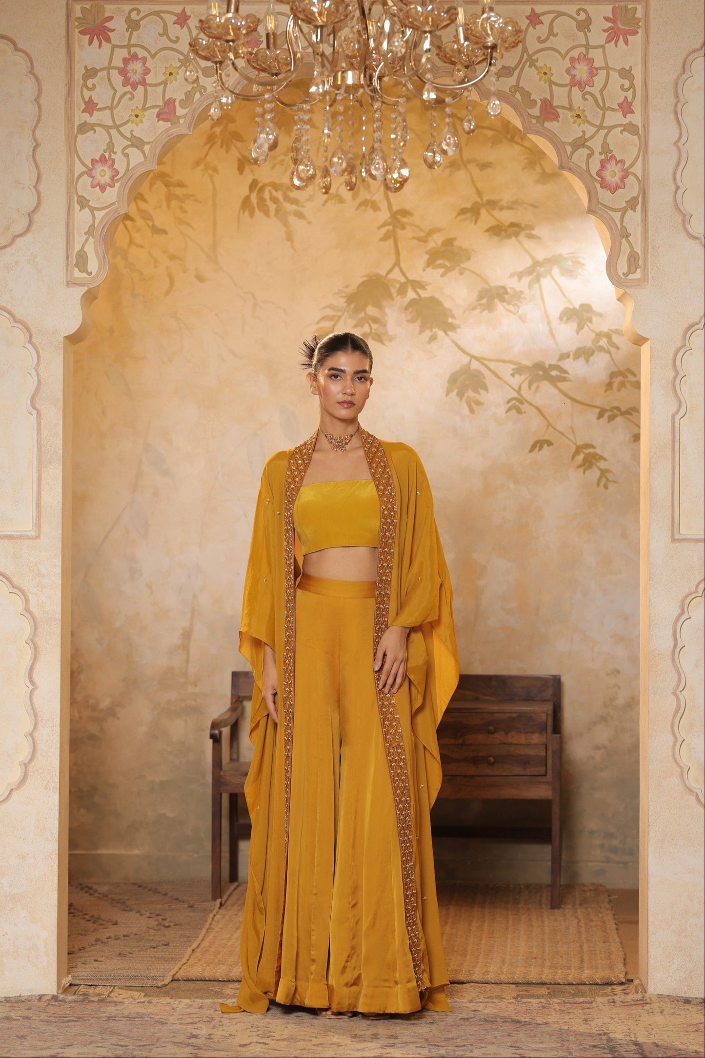 Woman in a mustard yellow outfit standing in an ornate room with a chandelier. Golden yellow sharara set, embroidered wedding outfit, designer festive wear, elegant ethnic ensemble, premium traditional attire, Sunanta Madaan