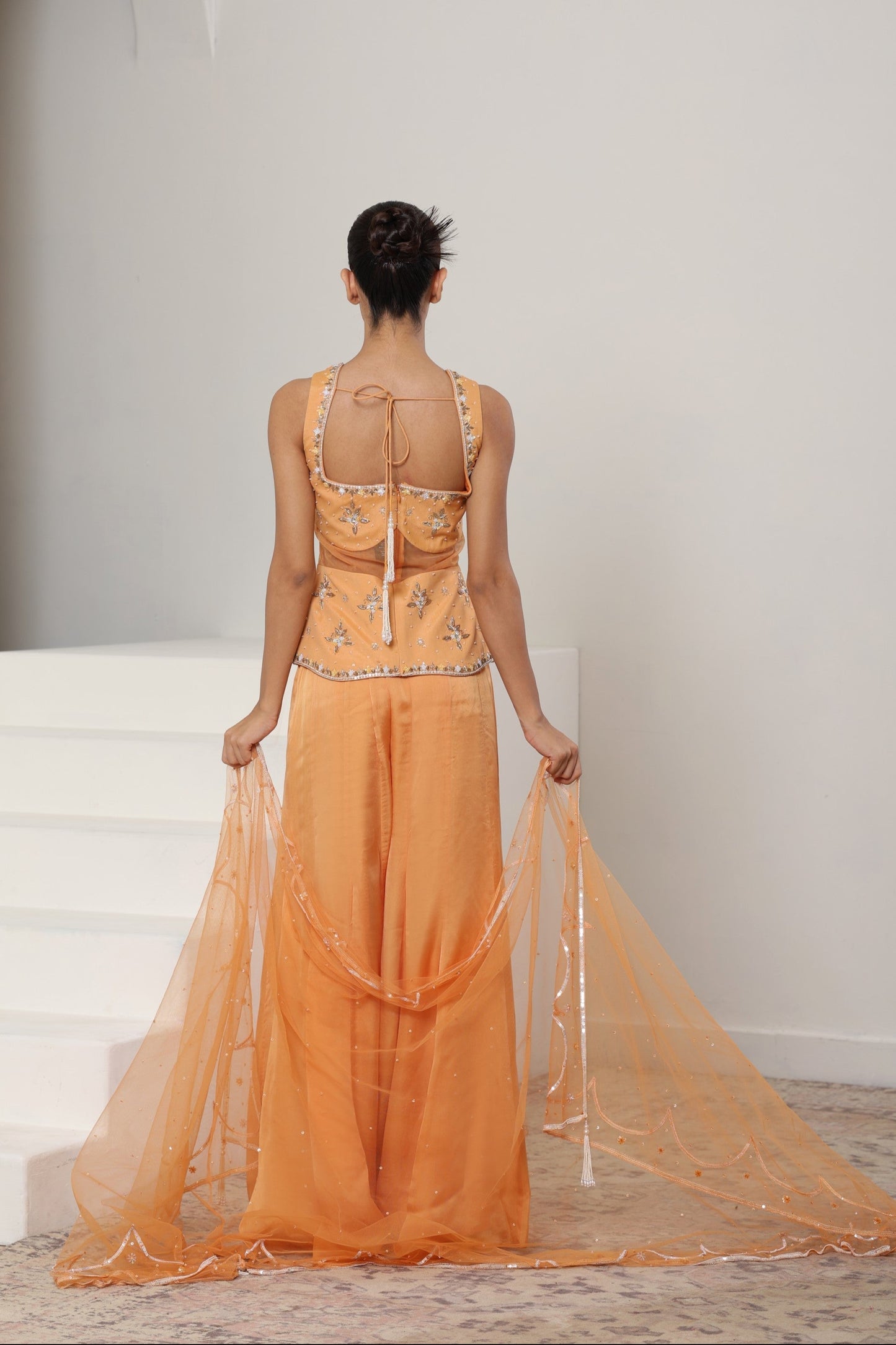 Woman wearing an orange dress with a sheer overlay in a minimal indoor setting; Orange silk crepe sharara, draped festive outfit, elegant ethnic wear, designer traditional attire, premium party ensemble, Sunanta Madaan, near me, Mumbai, Delhi, Hyderabad, Bangalore, Pune