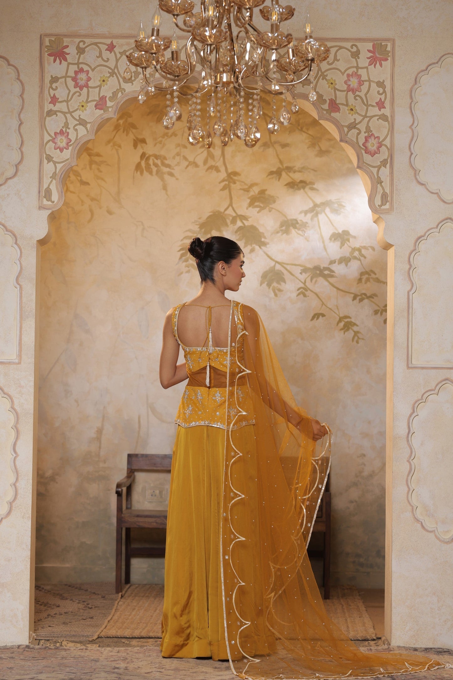 Woman in a yellow dress standing in a decorated room with a chandelier. Yellow silk crepe sharara, festive ethnic outfit, elegant occasional wear, designer traditional attire, premium party ensemble, Sunanta Madaan, near me, Mumbai, Delhi, Hyderabad, Bangalore, Pune