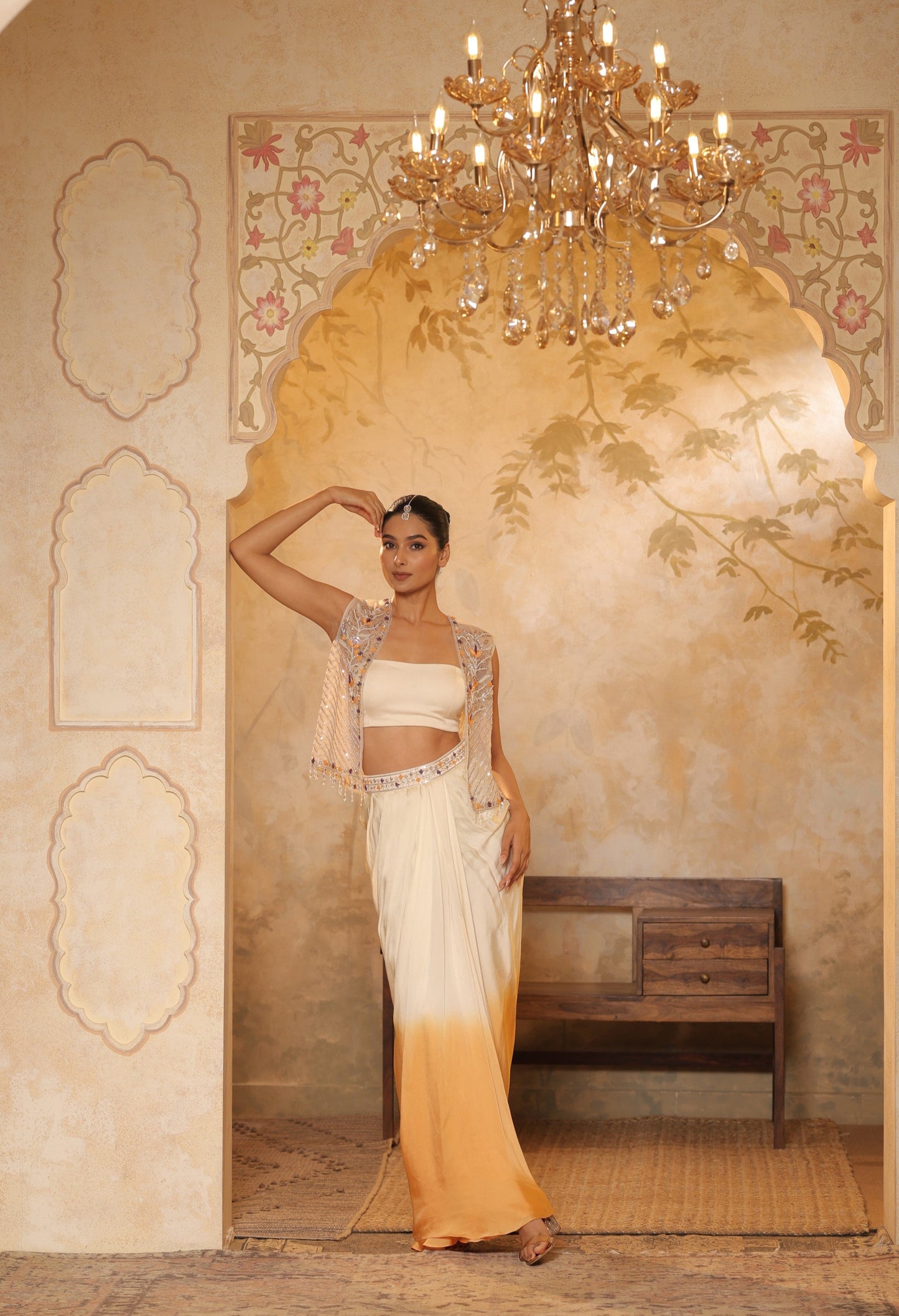 Woman in a traditional outfit standing in a room with a chandelier and decorative wall. Ivory orange skirt dress, hand embroidered crepe outfit, elegant festive wear, designer ethnic attire, premium traditional ensemble, Sunanta Madaan, near me, Mumbai, Delhi, Hyderabad, Bangalore, Pune