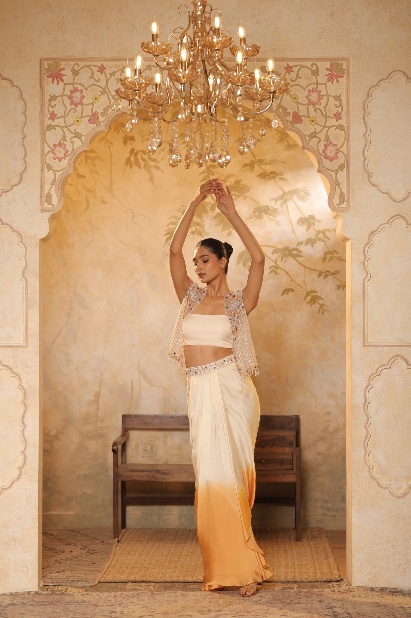 Woman in a traditional outfit standing in a decorated room with a chandelier. Ivory orange skirt dress, hand embroidered crepe outfit, elegant festive wear, designer ethnic attire, premium traditional ensemble, Sunanta Madaan, near me, Mumbai, Delhi, Hyderabad, Bangalore, Pune