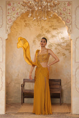 Woman in a mustard yellow traditional outfit standing in an ornate room with a chandelier. Golden yellow saree, chiffon embroidered saree, elegant traditional saree, festive ethnic wear, designer saree online, Sunanta Madaan