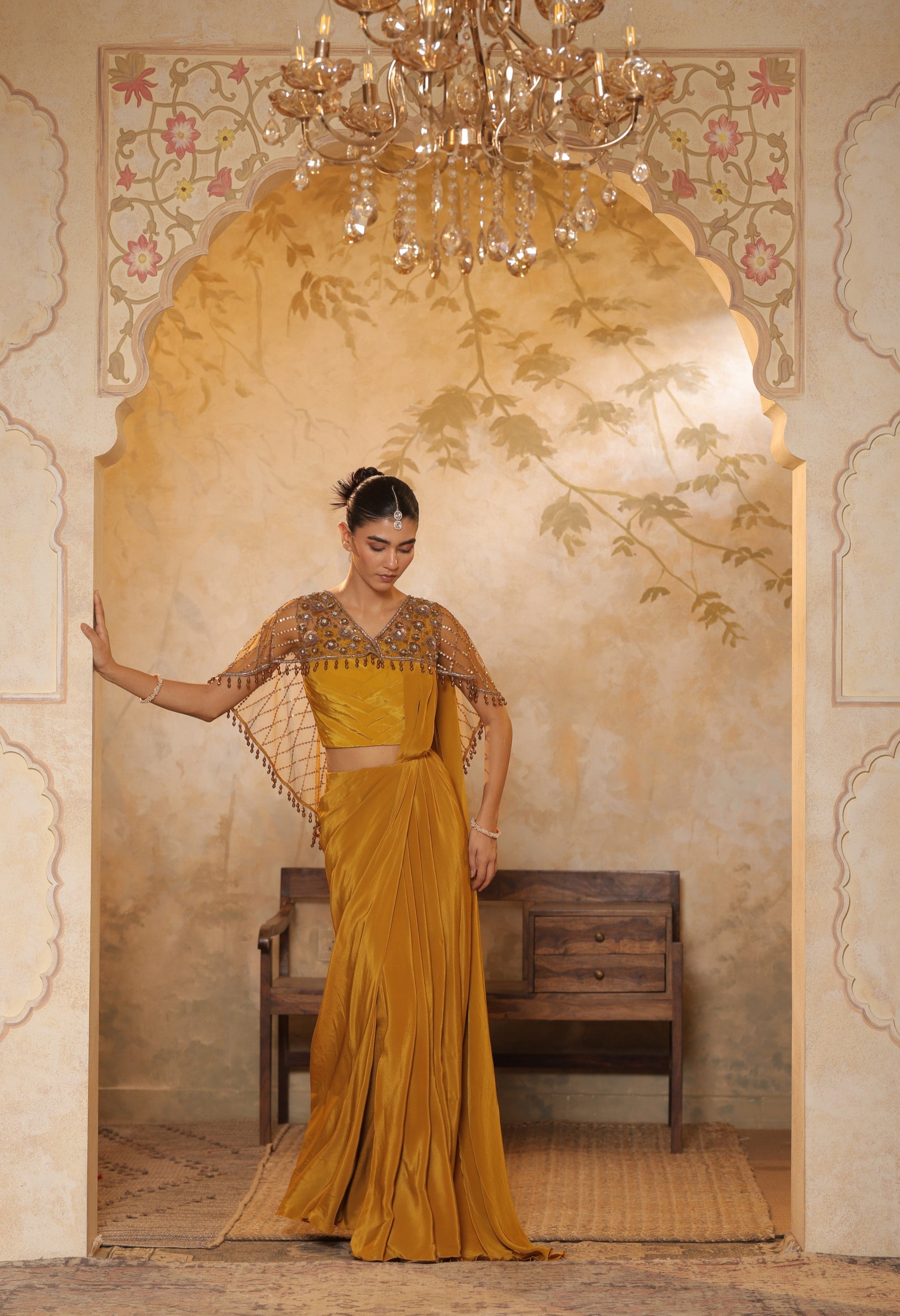Woman in a yellow saree with a decorative blouse standing in an ornate room. Golden yellow crepe saree, designer cape saree, premium haldi outfit, elegant festive wear, embroidered ethnic saree, Sunanta Madaan