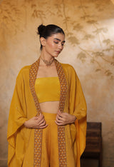Woman wearing a mustard yellow traditional outfit with intricate designs against a floral-patterned wall. Golden yellow sharara set, embroidered wedding outfit, designer festive wear, elegant ethnic ensemble, premium traditional attire, Sunanta Madaan
