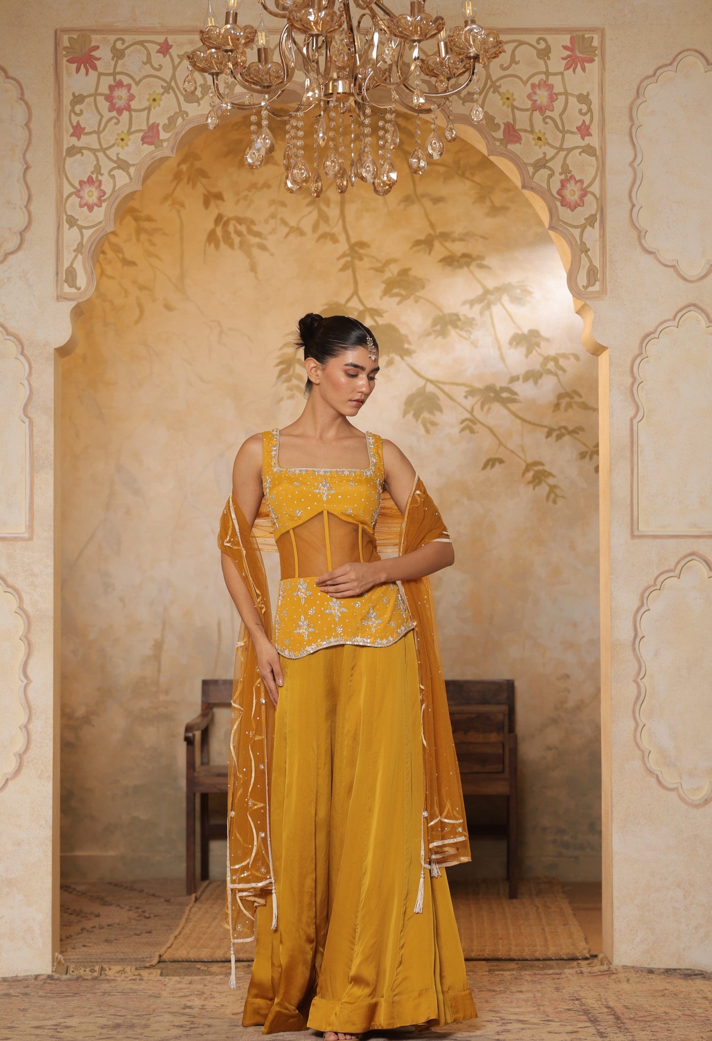 Woman in a yellow traditional outfit standing in an ornate room with a chandelier. Yellow silk crepe sharara, festive ethnic outfit, elegant occasional wear, designer traditional attire, premium party ensemble, Sunanta Madaan, near me, Mumbai, Delhi, Hyderabad, Bangalore, Pune