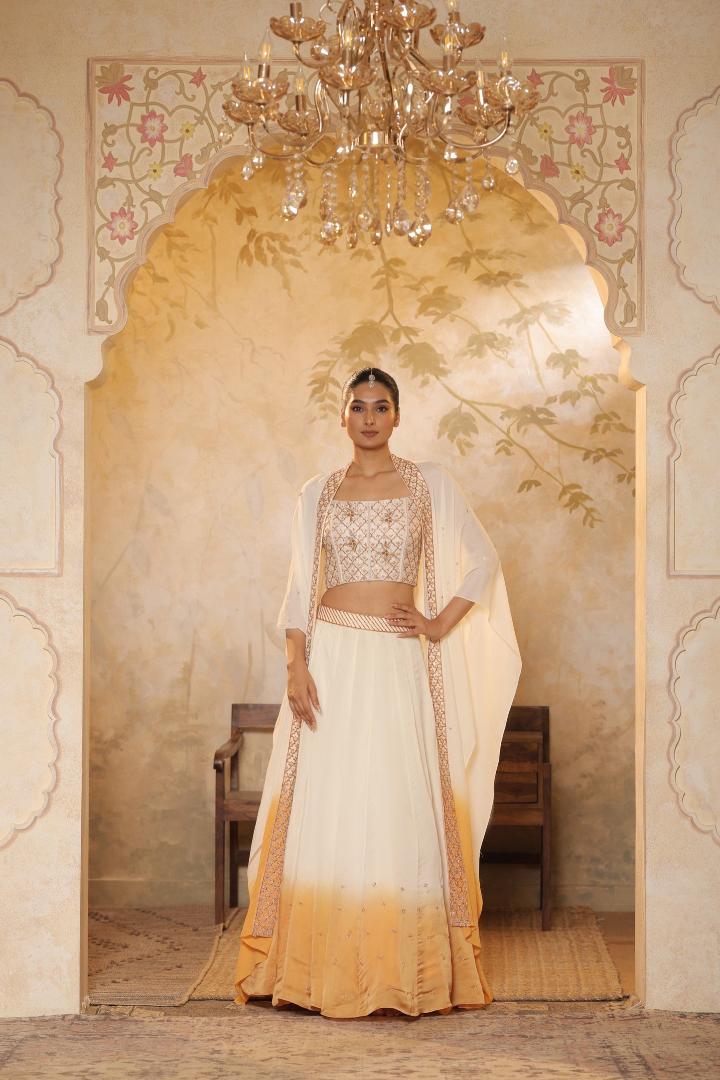 Woman in a traditional outfit standing in an ornate doorway with a chandelier above. Ivory orange organza lehenga, satin embroidered lehenga set, festive lehenga with cape, elegant designer outfit, ethnic party wear, Sunanta Madaan