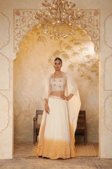 Woman in a traditional outfit standing in an ornate doorway with a chandelier above. Ivory orange organza lehenga, satin embroidered lehenga set, festive lehenga with cape, elegant designer outfit, ethnic party wear, Sunanta Madaan