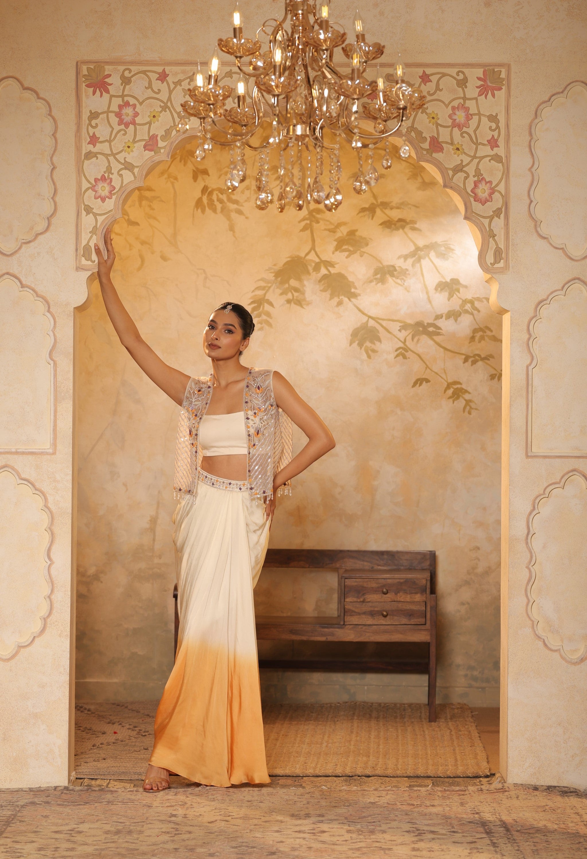 Woman in traditional outfit standing in a decorated room with a chandelier. Ivory orange skirt dress, hand embroidered crepe outfit, elegant festive wear, designer ethnic attire, premium traditional ensemble, Sunanta Madaan, near me, Mumbai, Delhi, Hyderabad, Bangalore, Pune