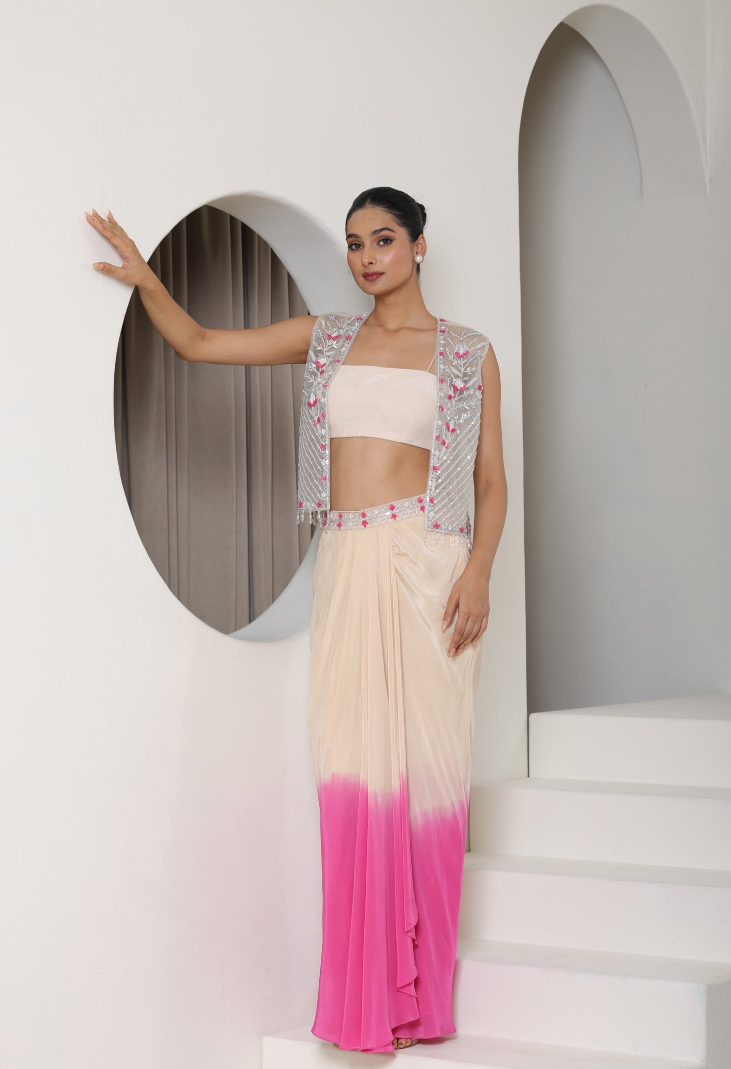 Woman in a pink and white outfit with a gradient skirt, standing against a white wall with an arch. Ivory pink skirt dress, viscose crepe outfit, pre-draped designer wear, premium occasion attire, elegant festive ensemble, Sunanta Madaan, near me, Mumbai, Delhi, Hyderabad, Bangalore, Pune