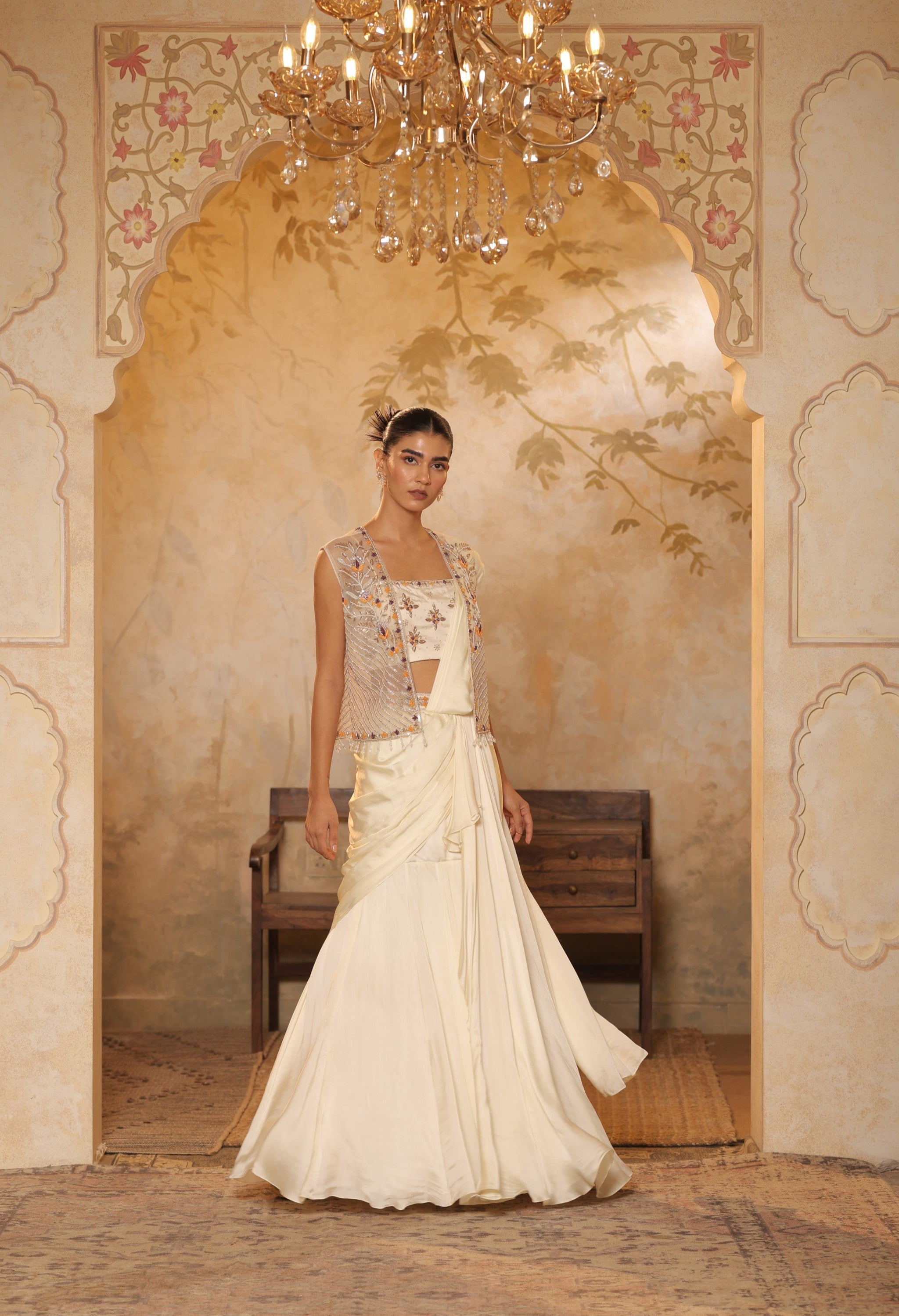 Woman in a traditional outfit standing in an ornate room with a chandelier. ivory georgette saree, saree with embroidered cape, designer wedding saree India, Indo Western bridal outfit, elegant ivory saree, Sunanta Madaan