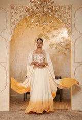 Woman in a traditional outfit with a yellow and white dress in an ornate room. Ivory orange organza lehenga, satin embroidered lehenga set, festive lehenga with cape, elegant designer outfit, ethnic party wear, Sunanta Madaan