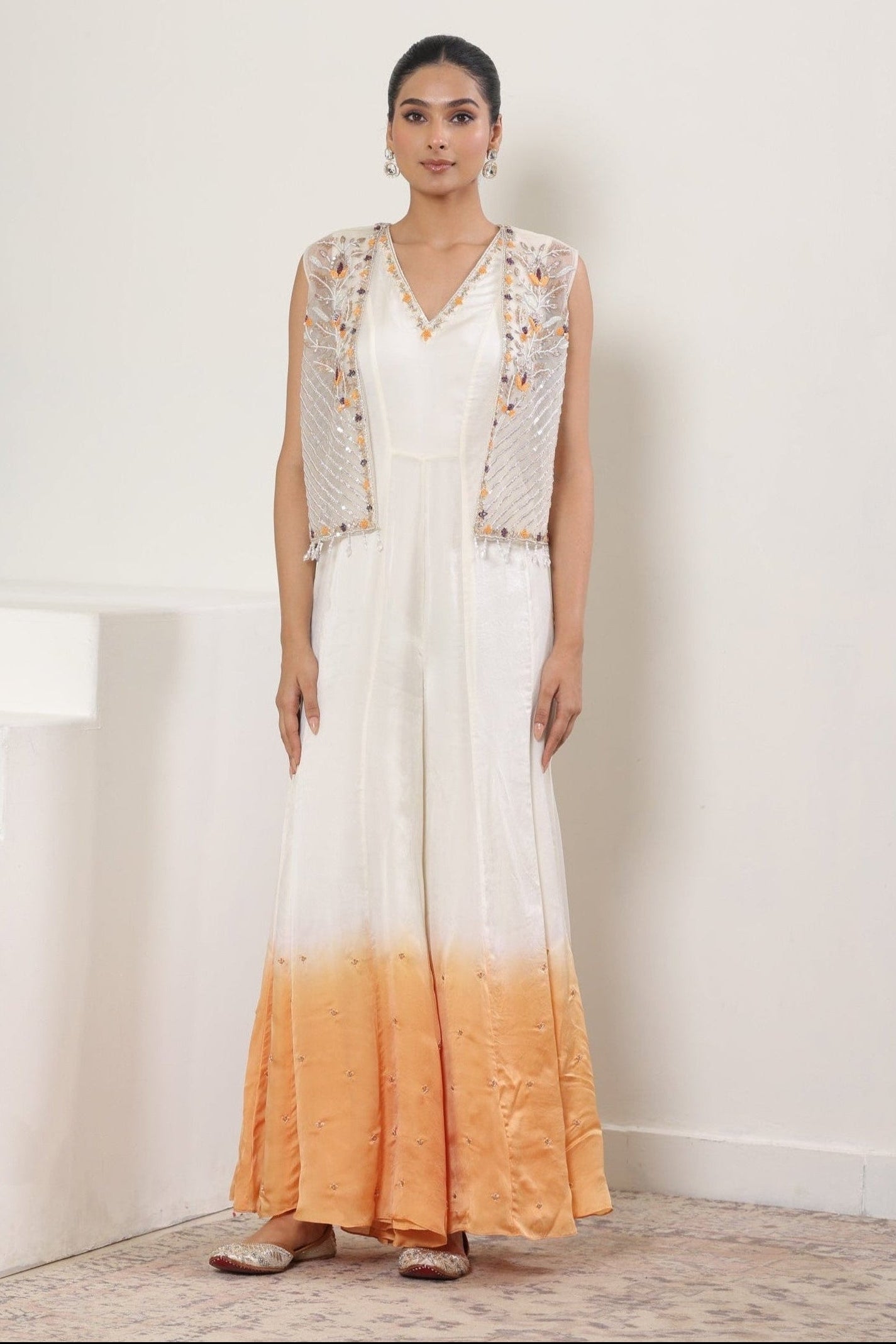 Woman wearing a white and orange dress with a matching jacket on a plain background; Ivory orange jumpsuit, satin organza outfit, designer cape jumpsuit, elegant occasional wear, festive ethnic attire, Sunanta Madaan, near me, Mumbai, Delhi, Hyderabad, Bangalore, Pune