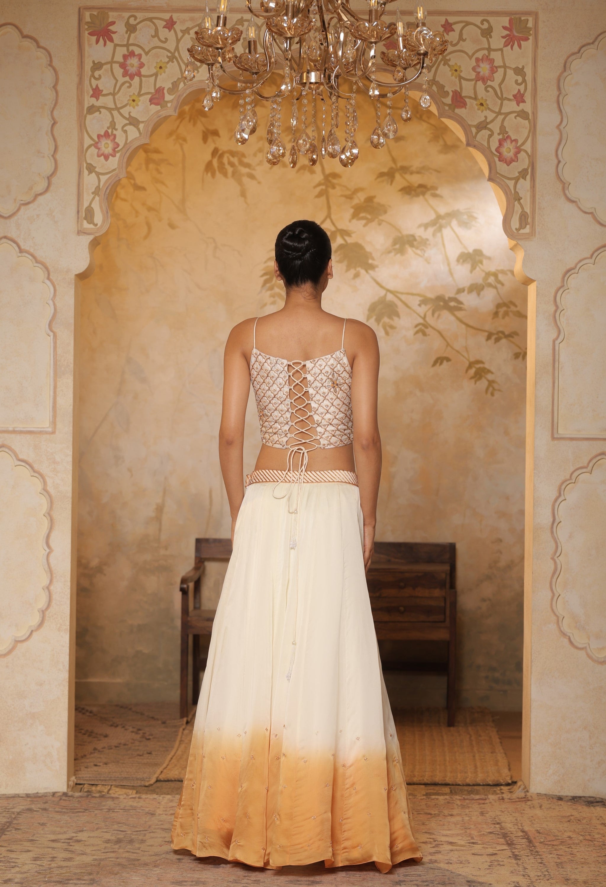 Woman wearing a white and gold outfit in an ornate room with a chandelier. Ivory orange organza lehenga, satin embroidered lehenga set, festive lehenga with cape, elegant designer outfit, ethnic party wear, Sunanta Madaan