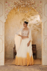 Woman in a traditional outfit standing in an ornate room with a chandelier.Ivory orange organza lehenga, satin embroidered lehenga set, festive lehenga with cape, elegant designer outfit, ethnic party wear, Sunanta Madaan