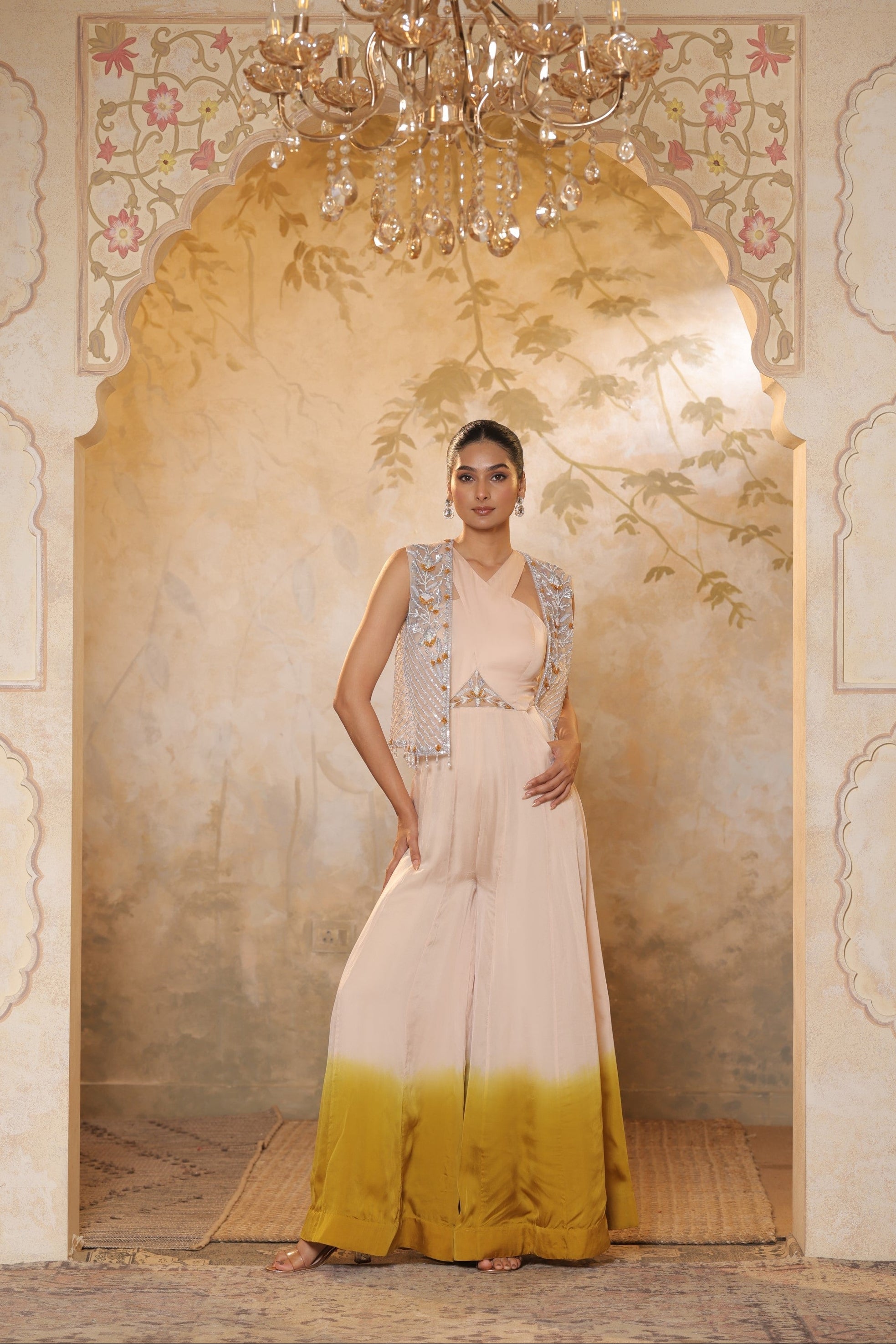 Woman in a white and yellow dress standing in an ornate doorway with a chandelier above. Sunanta Madaan, Ivory yellow jumpsuit, satin organza outfit, sequin work haldi wear, premium festive attire, elegant designer jumpsuit, Mumbai, Delhi, Hyderabad, Bangalore, Pune
