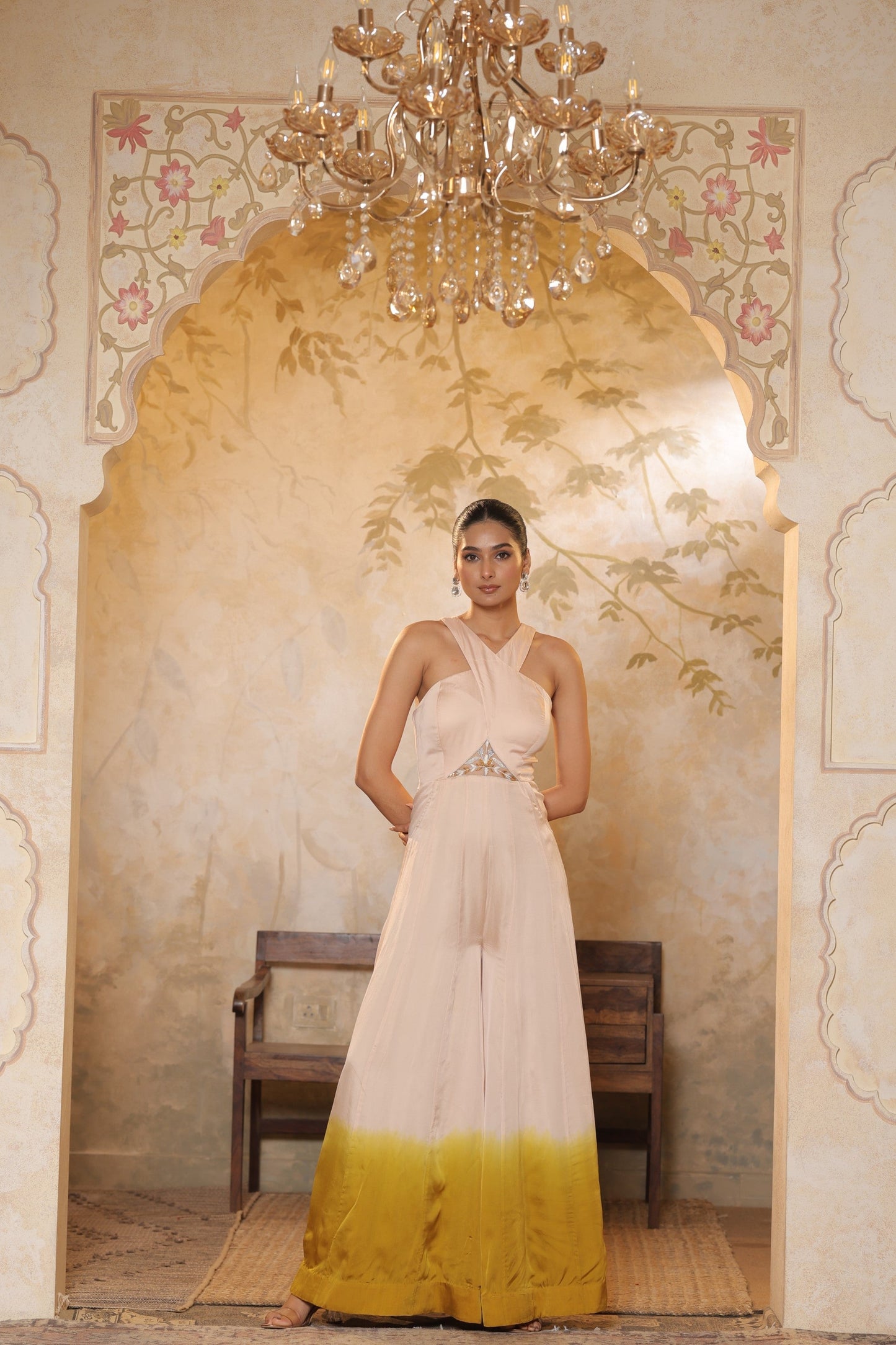 Woman in a white and yellow dress standing in an ornate room with a chandelier. Ivory yellow jumpsuit, satin organza outfit, sequin work haldi wear, premium festive attire, elegant designer jumpsuit, Sunanta Madaan