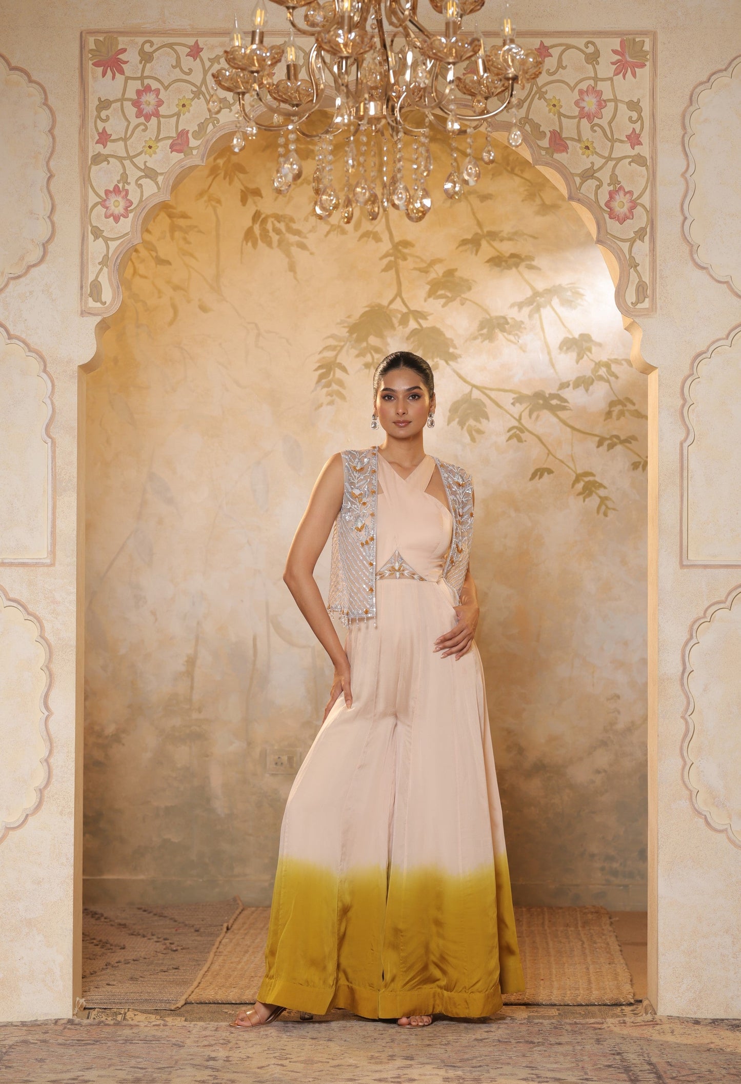 Woman in a white and yellow dress standing in an ornate doorway with a chandelier above. Sunanta Madaan, Ivory yellow jumpsuit, satin organza outfit, sequin work haldi wear, premium festive attire, elegant designer jumpsuit, Mumbai, Delhi, Hyderabad, Bangalore, Pune