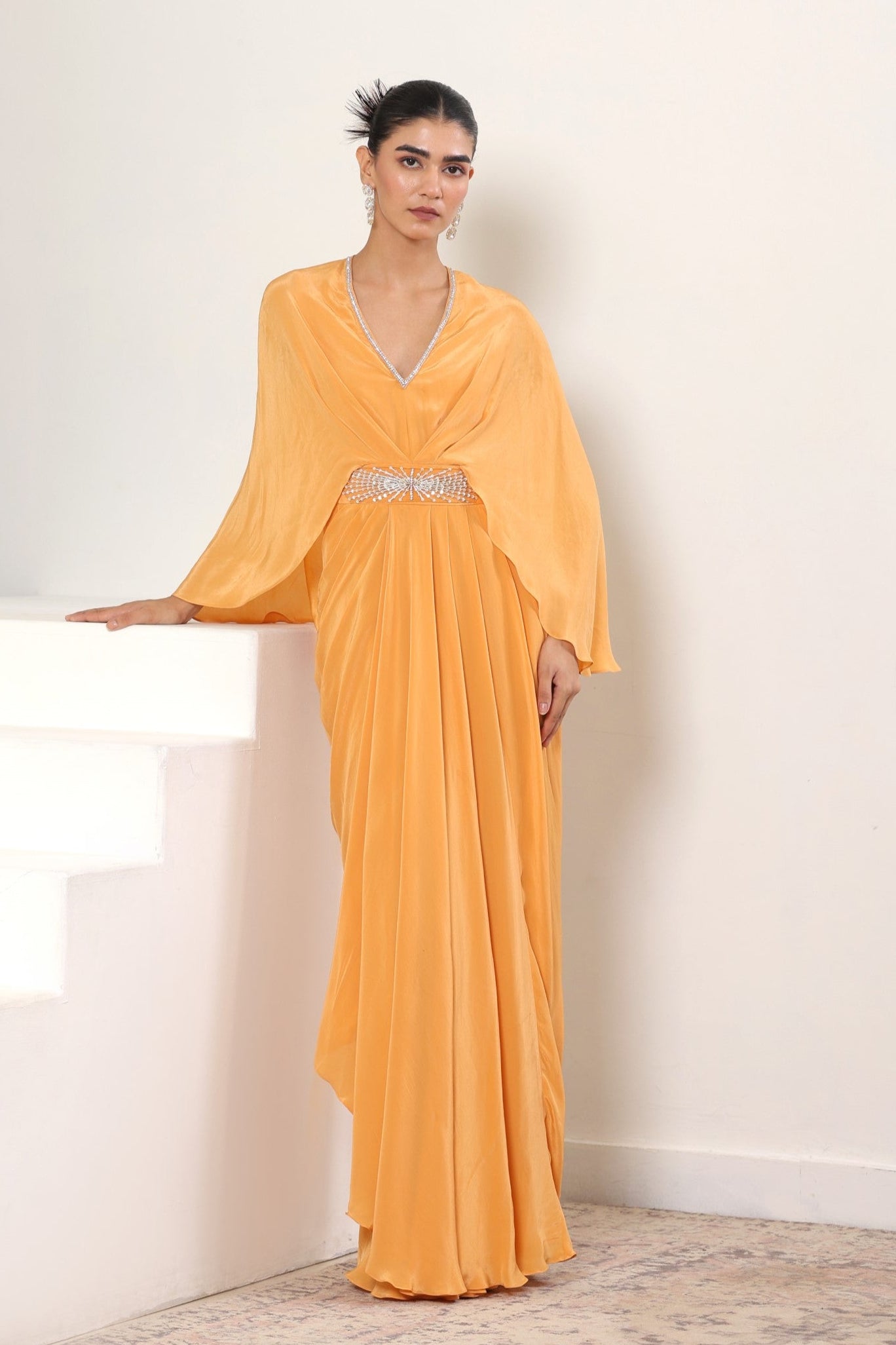 Woman wearing a long orange dress with a V-neckline and embellishments on a white background; Orange crepe kaftan, embroidered designer kaftan, luxury evening wear, elegant festive outfit, premium ethnic kaftan, Sunanta Madaan