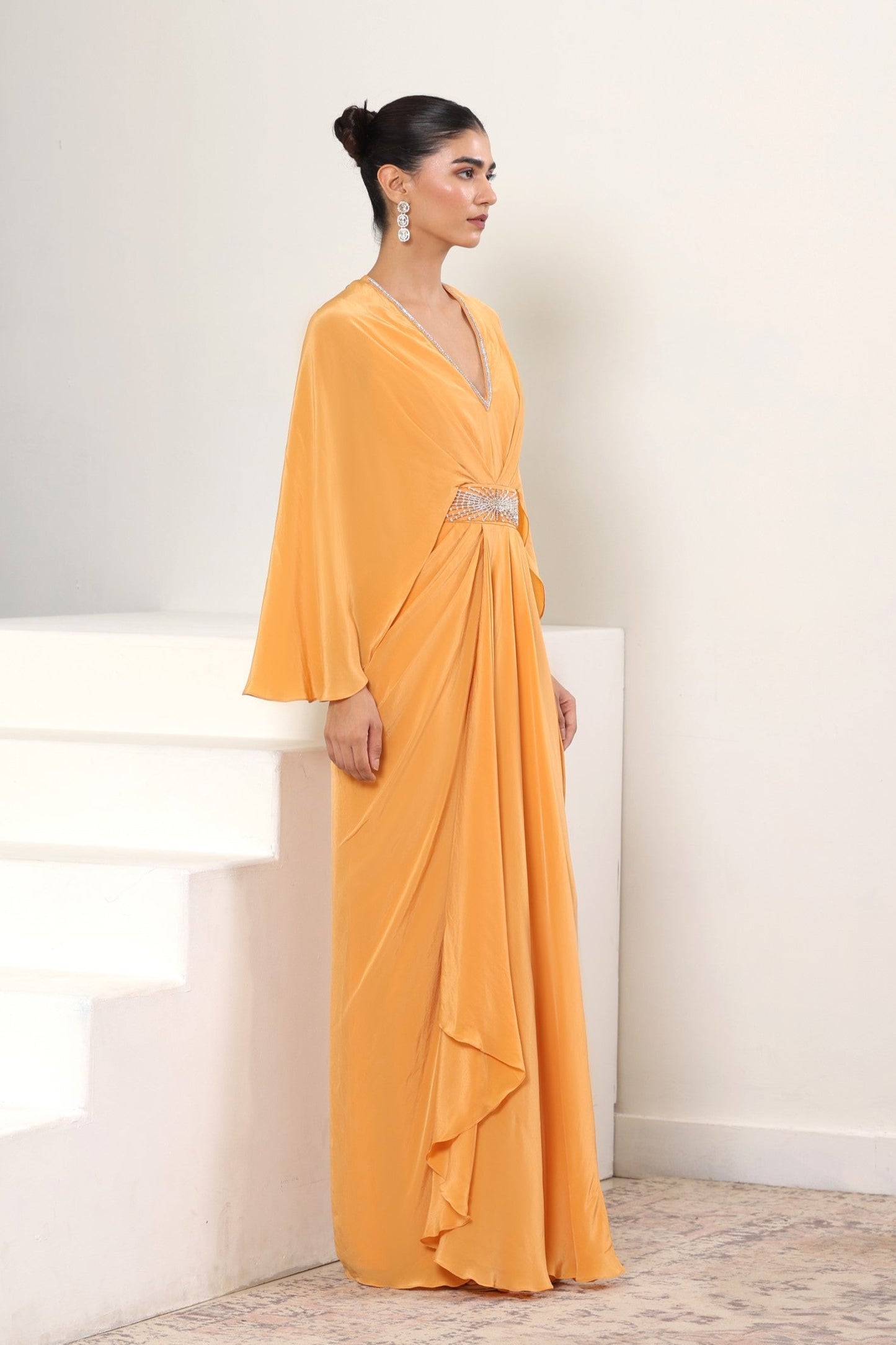 Woman in a long orange dress standing on a staircase with a white wall background; Orange crepe kaftan, embroidered designer kaftan, luxury evening wear, elegant festive outfit, premium ethnic kaftan, Sunanta Madaan