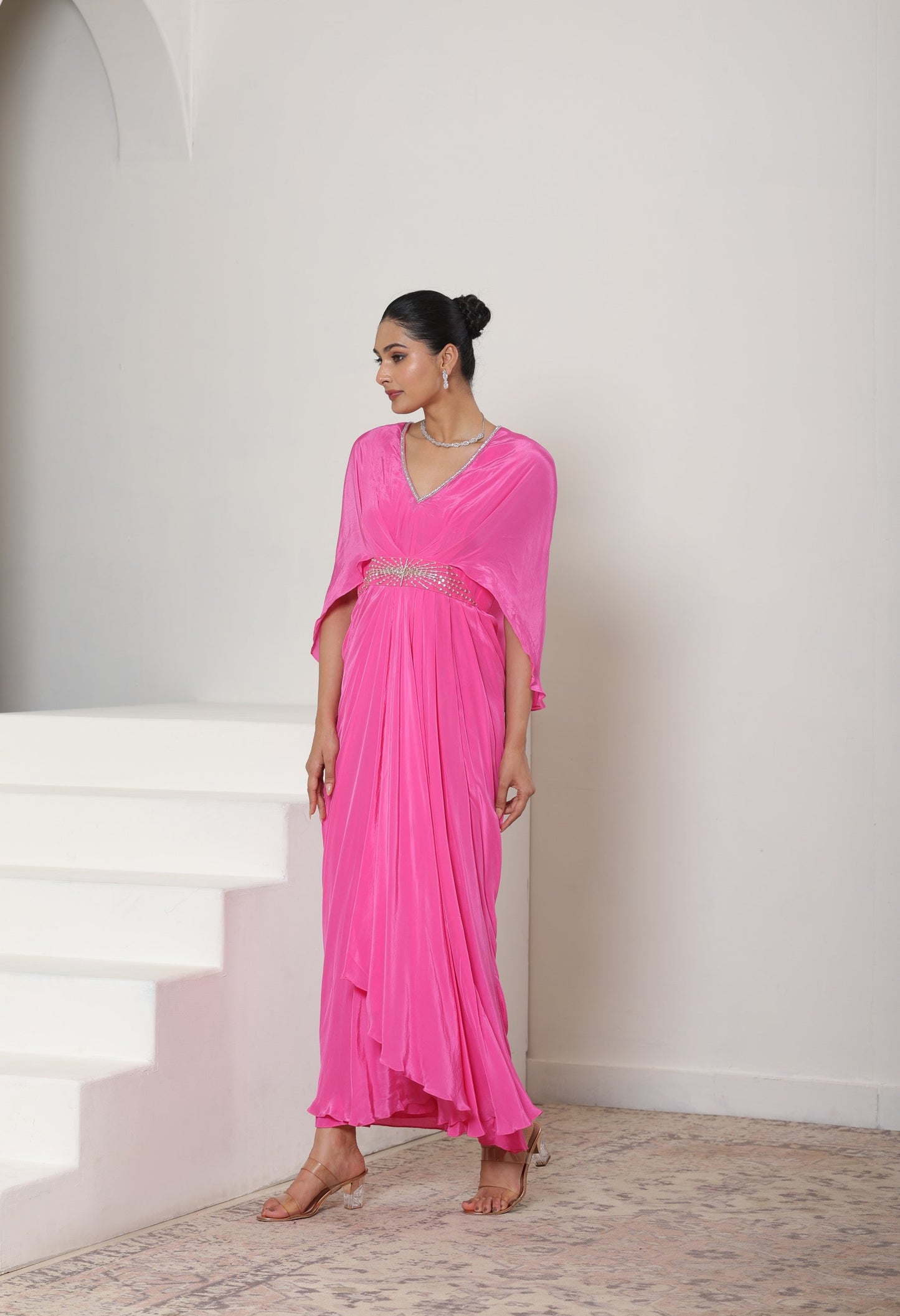 Woman in a pink outfit standing on a staircase with a white wall background; Pink crepe kaftan, V-neck designer kaftan, elegant cocktail outfit, premium evening wear, stylish festive kaftan, Sunanta Madaan