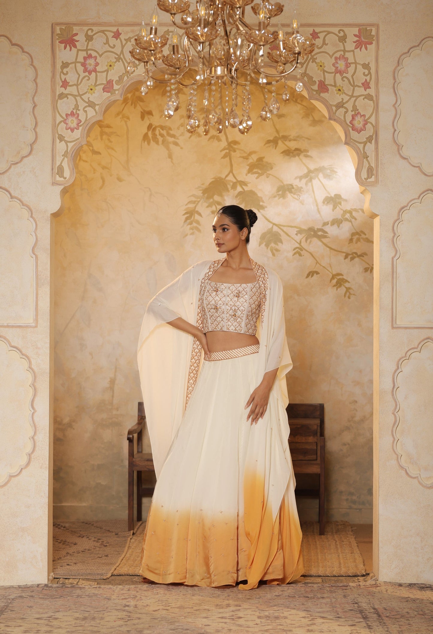 Woman in a traditional outfit standing in an ornate room with a chandelier.Ivory orange organza lehenga, satin embroidered lehenga set, festive lehenga with cape, elegant designer outfit, ethnic party wear, Sunanta Madaan