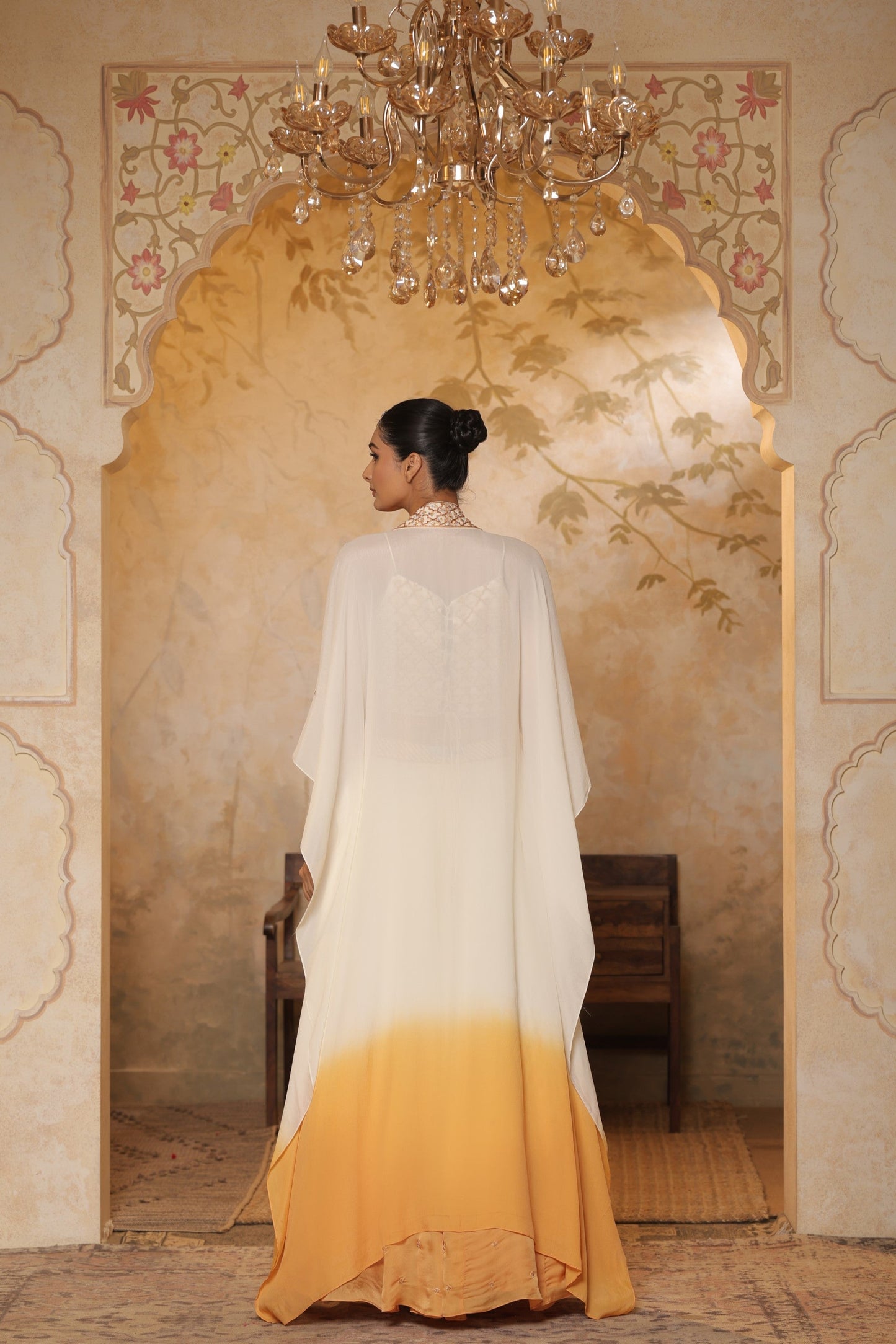 Woman in a white and yellow dress standing in a decorated room with a chandelier. Ivory orange organza lehenga, satin embroidered lehenga set, festive lehenga with cape, elegant designer outfit, ethnic party wear, Sunanta Madaan