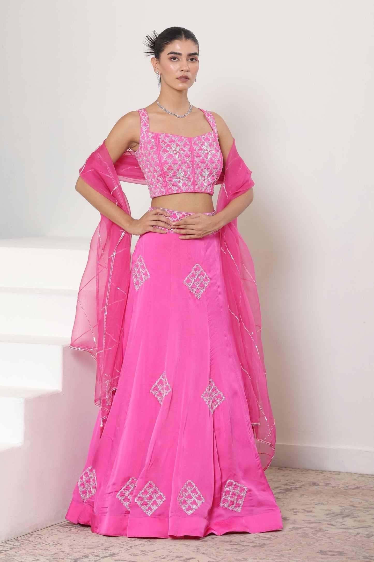 Woman wearing a pink traditional outfit with white patterns on a light background; Pink satin organza lehenga, embroidered designer outfit, premium reception wear, elegant festive attire, traditional bridal ensemble, Sunanta Madaan, Boho Indo-Western dress, Indo-Western fusion wear, Indo-Western attire for engagement, Indo-Western outfit for sangeet, Indo-Western draped dress, Indo-Western embroidered skirt, Indo-Western embroidered dress, near me, Mumbai, Delhi, Hyderabad, Bangalore, Pune