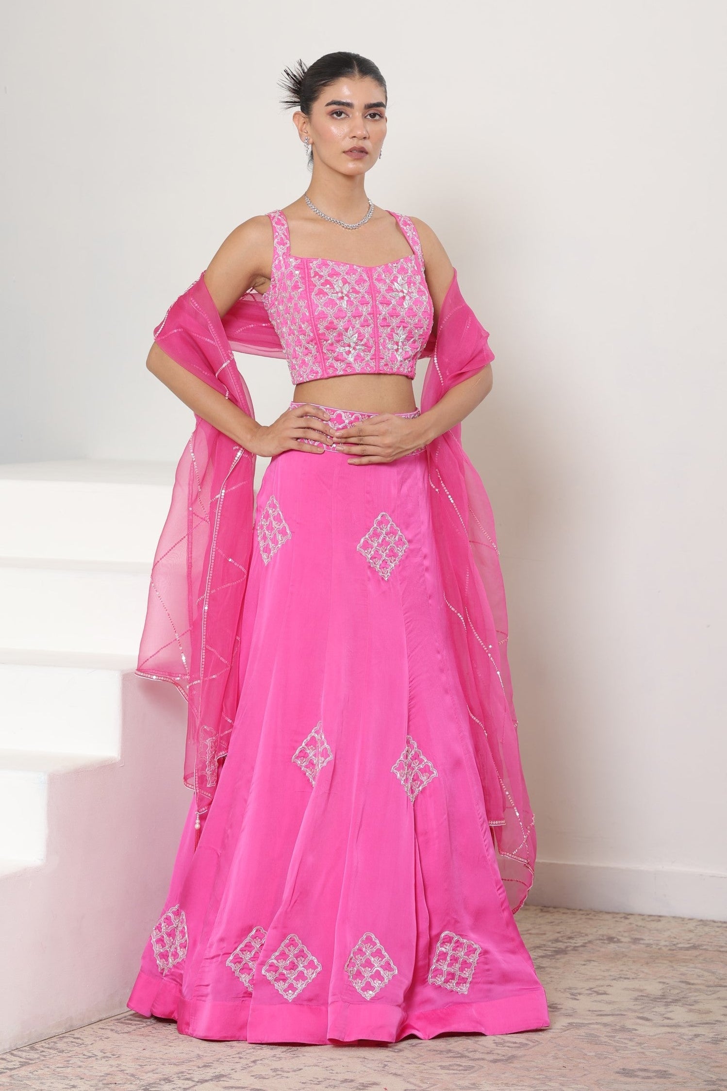Woman wearing a pink traditional outfit with white patterns on a light background; Pink satin organza lehenga, embroidered designer outfit, premium reception wear, elegant festive attire, traditional bridal ensemble, Sunanta Madaan, Boho Indo-Western dress, Indo-Western fusion wear, Indo-Western attire for engagement, Indo-Western outfit for sangeet, Indo-Western draped dress, Indo-Western embroidered skirt, Indo-Western embroidered dress, near me, Mumbai, Delhi, Hyderabad, Bangalore, Pune