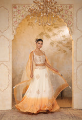 Woman in a white and orange traditional outfit standing in a decorated room with a chandelier. Sunanta Madaan – Ivory-Orange Satin Organza Lehenga, Embroidered Designer Outfit, Elegant Festive Attire, Premium Party & Reception Wear, Boho Indo-Western Dress, Indo-Western Fusion Wear, Indo-Western Attire for Engagement, Indo-Western Outfit for Sangeet, Indo-Western Draped & Embroidered Skirt, Mumbai, Delhi, Hyderabad, Bangalore, Pune, near me