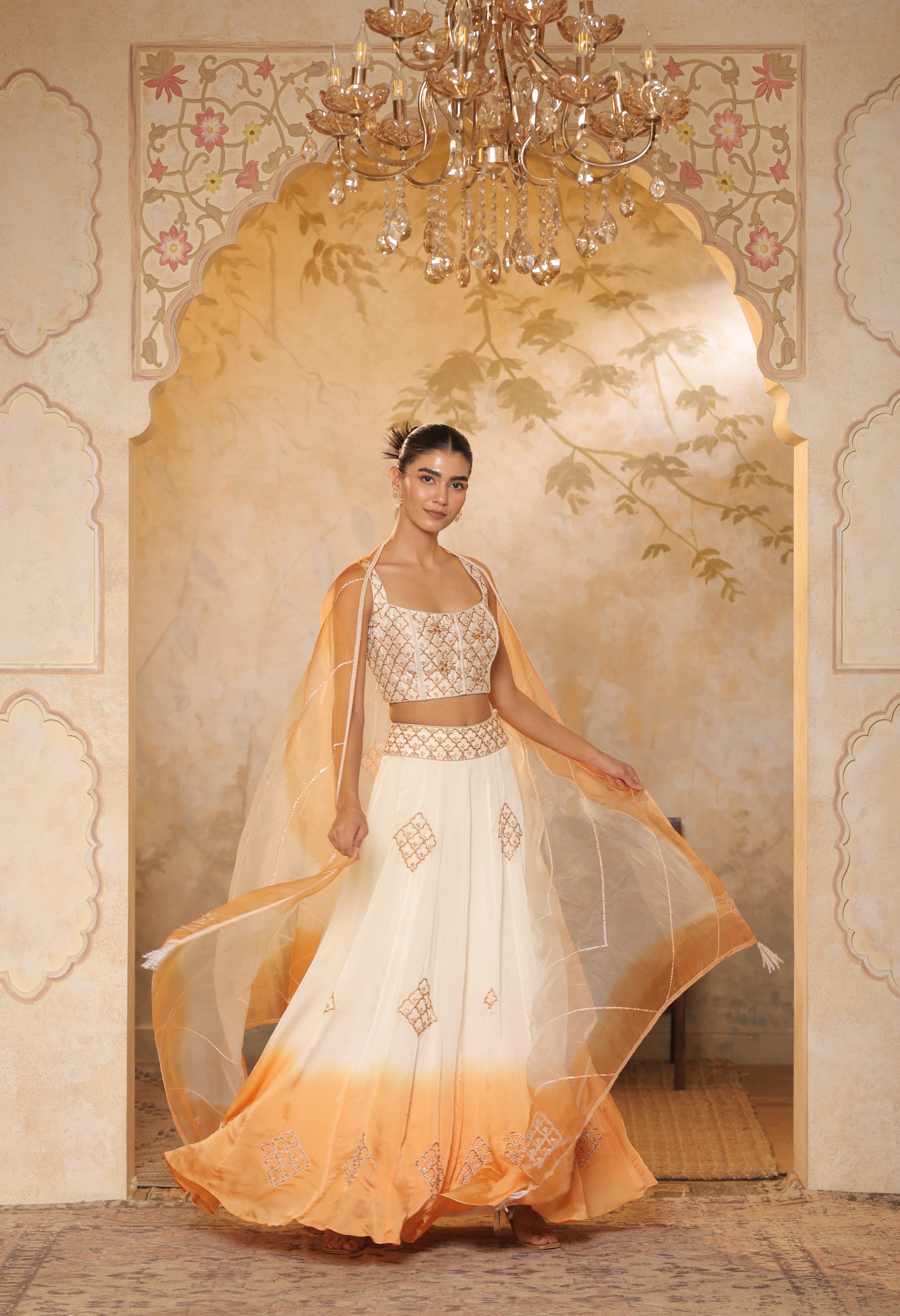 Woman in a white and orange traditional outfit standing in a decorated room with a chandelier. Sunanta Madaan – Ivory-Orange Satin Organza Lehenga, Embroidered Designer Outfit, Elegant Festive Attire, Premium Party & Reception Wear, Boho Indo-Western Dress, Indo-Western Fusion Wear, Indo-Western Attire for Engagement, Indo-Western Outfit for Sangeet, Indo-Western Draped & Embroidered Skirt, Mumbai, Delhi, Hyderabad, Bangalore, Pune, near me