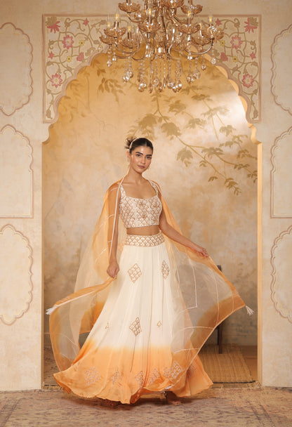 Woman in a white and orange traditional outfit standing in a decorated room with a chandelier. Sunanta Madaan – Ivory-Orange Satin Organza Lehenga, Embroidered Designer Outfit, Elegant Festive Attire, Premium Party & Reception Wear, Boho Indo-Western Dress, Indo-Western Fusion Wear, Indo-Western Attire for Engagement, Indo-Western Outfit for Sangeet, Indo-Western Draped & Embroidered Skirt, Mumbai, Delhi, Hyderabad, Bangalore, Pune, near me