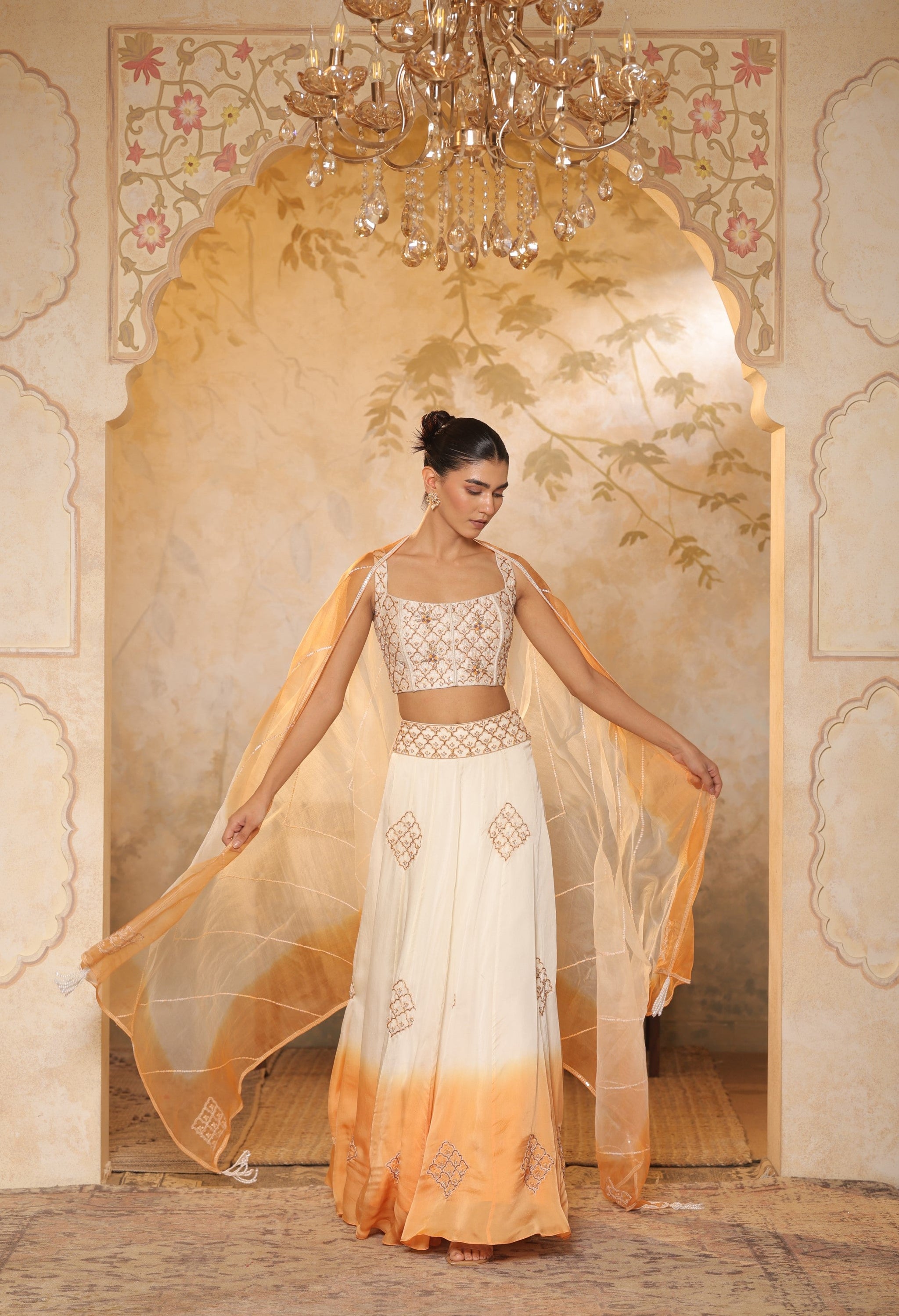 Woman in a white and orange traditional outfit standing in a decorated room with a chandelier. Sunanta Madaan – Ivory-Orange Satin Organza Lehenga, Embroidered Designer Outfit, Elegant Festive Attire, Premium Party & Reception Wear, Boho Indo-Western Dress, Indo-Western Fusion Wear, Indo-Western Attire for Engagement, Indo-Western Outfit for Sangeet, Indo-Western Draped & Embroidered Skirt, Mumbai, Delhi, Hyderabad, Bangalore, Pune, near me